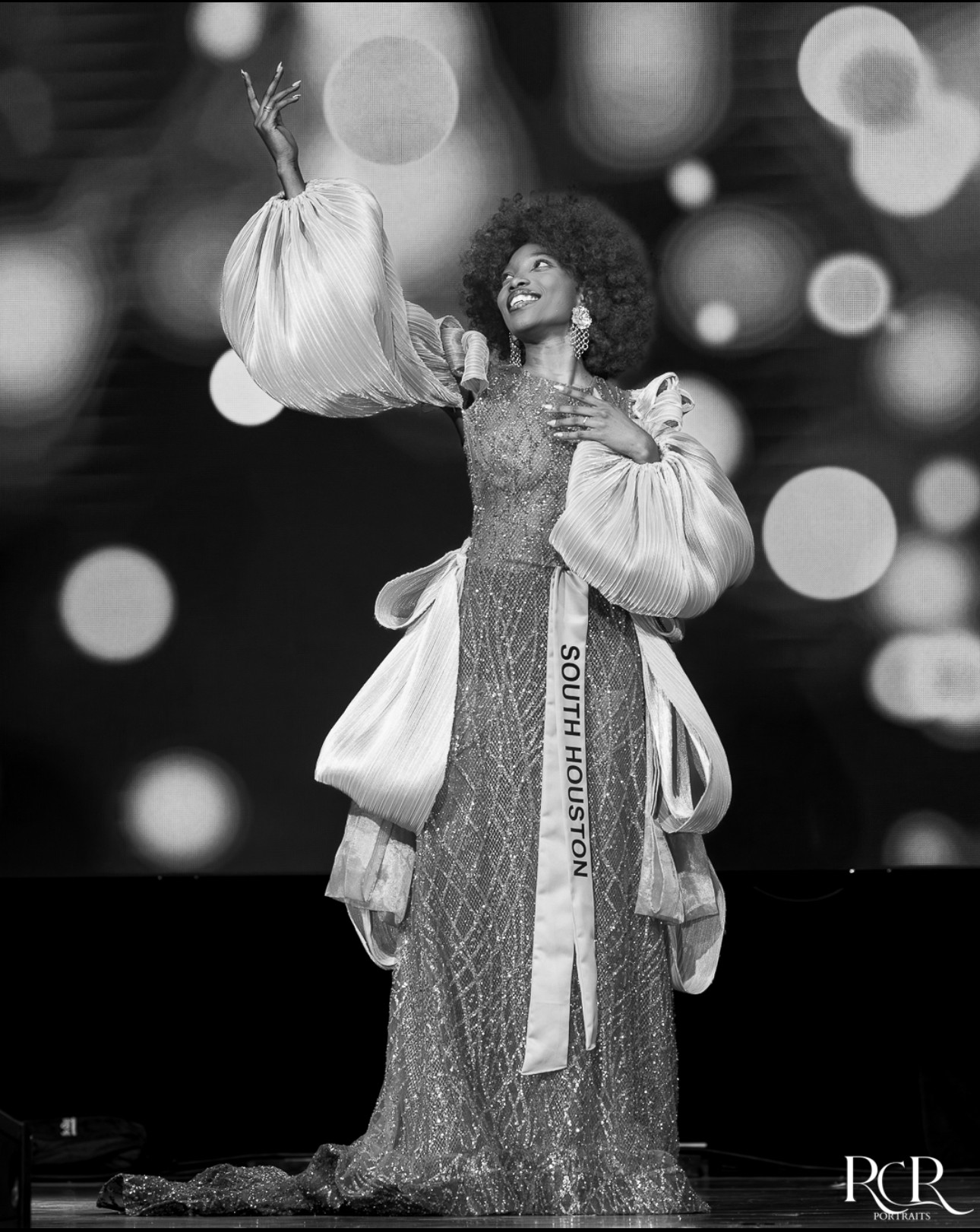 Woman in elaborate dress holding a large bag, smiling, with blurred circular lights in background.
