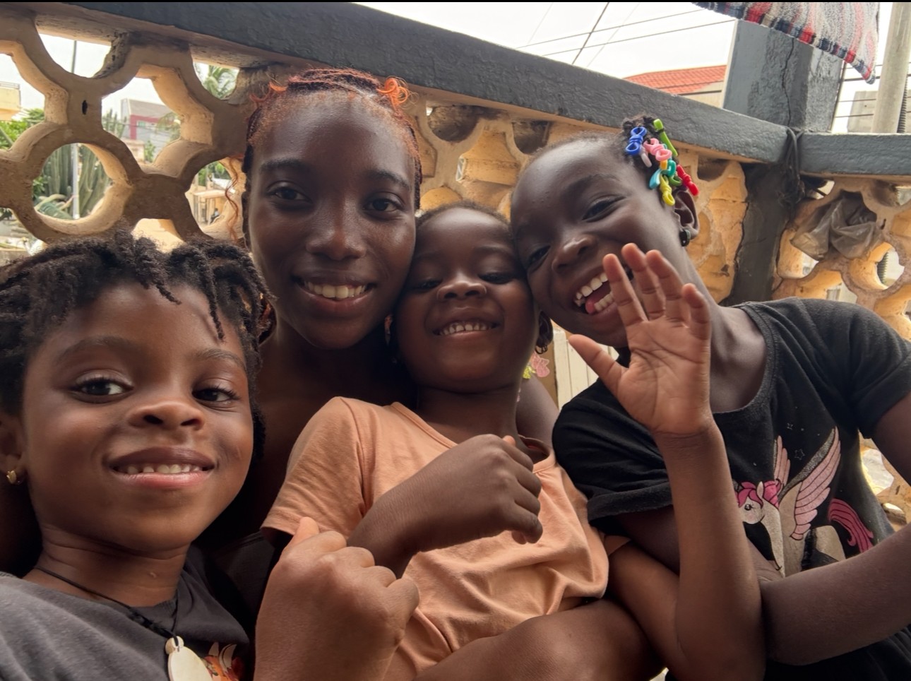 Four children smiling and posing together outdoors near a decorative wall.