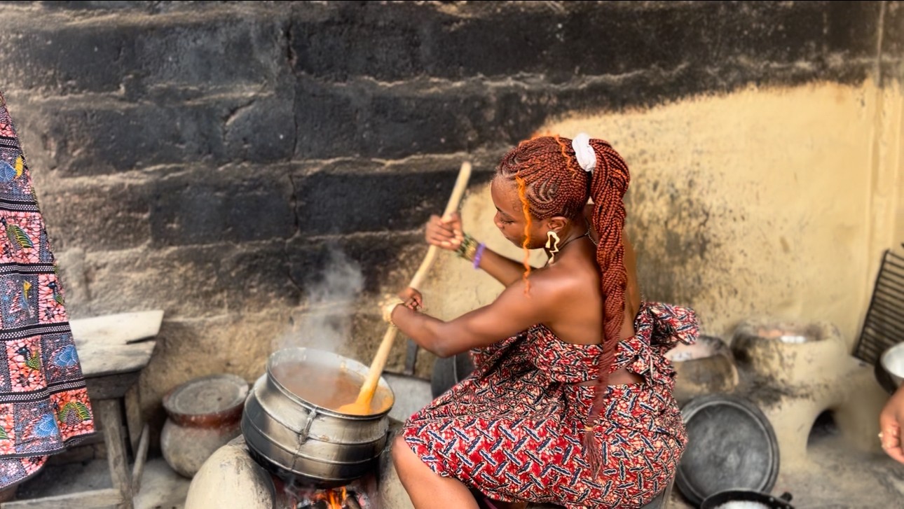 Woman cooking outdoors with a large pot, stirring with a wooden stick, against a brick wall background.