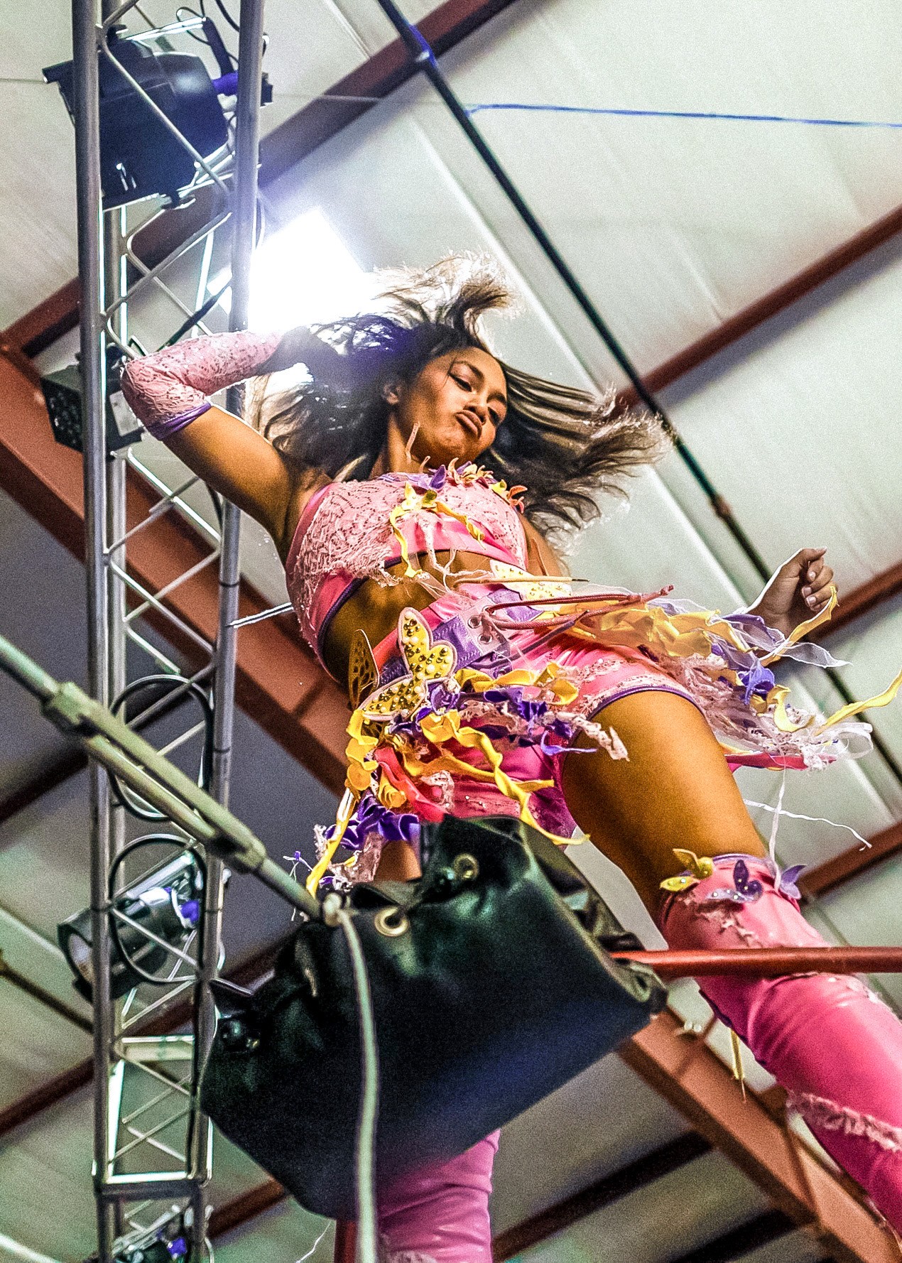 Woman in colorful outfit lying on scaffolding, hair flowing, with a black bag hanging nearby.