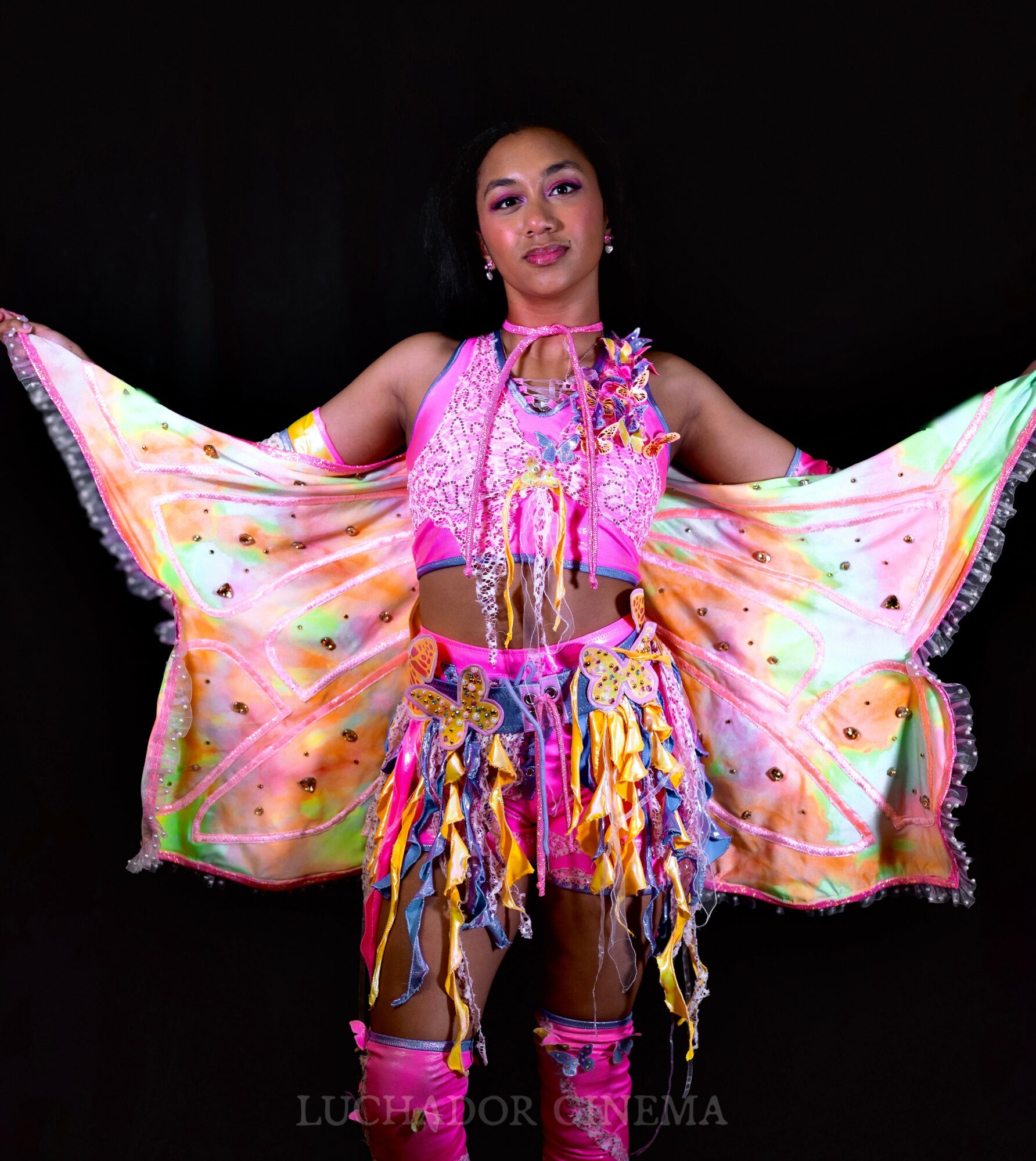 Young woman in colorful costume with wings, holding fabric outstretched, against dark background.