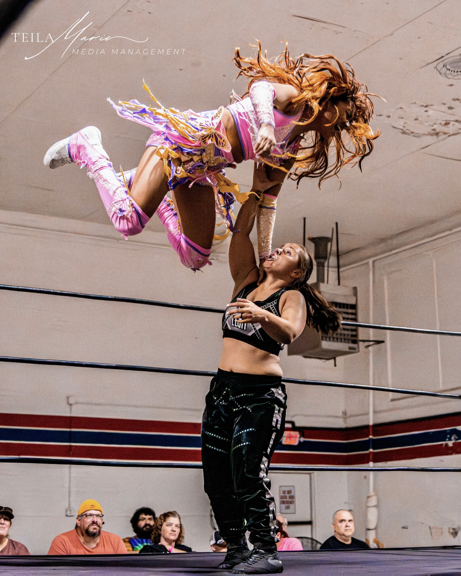 Two women perform a dance lift in a gym, with spectators watching in the background.