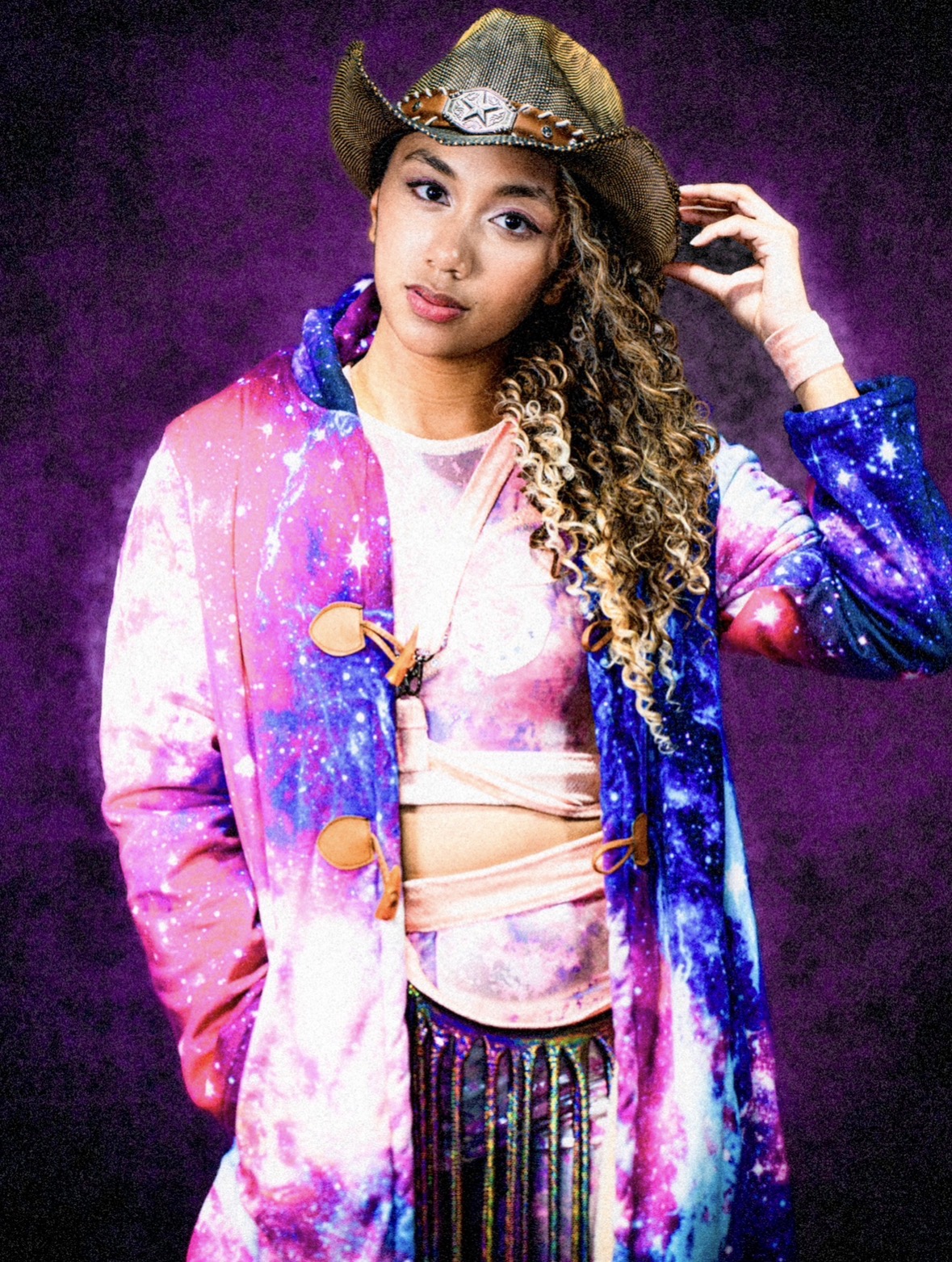 Young woman with curly hair wearing a galaxy-themed jacket, hat, and colorful clothing, standing against a purple background.