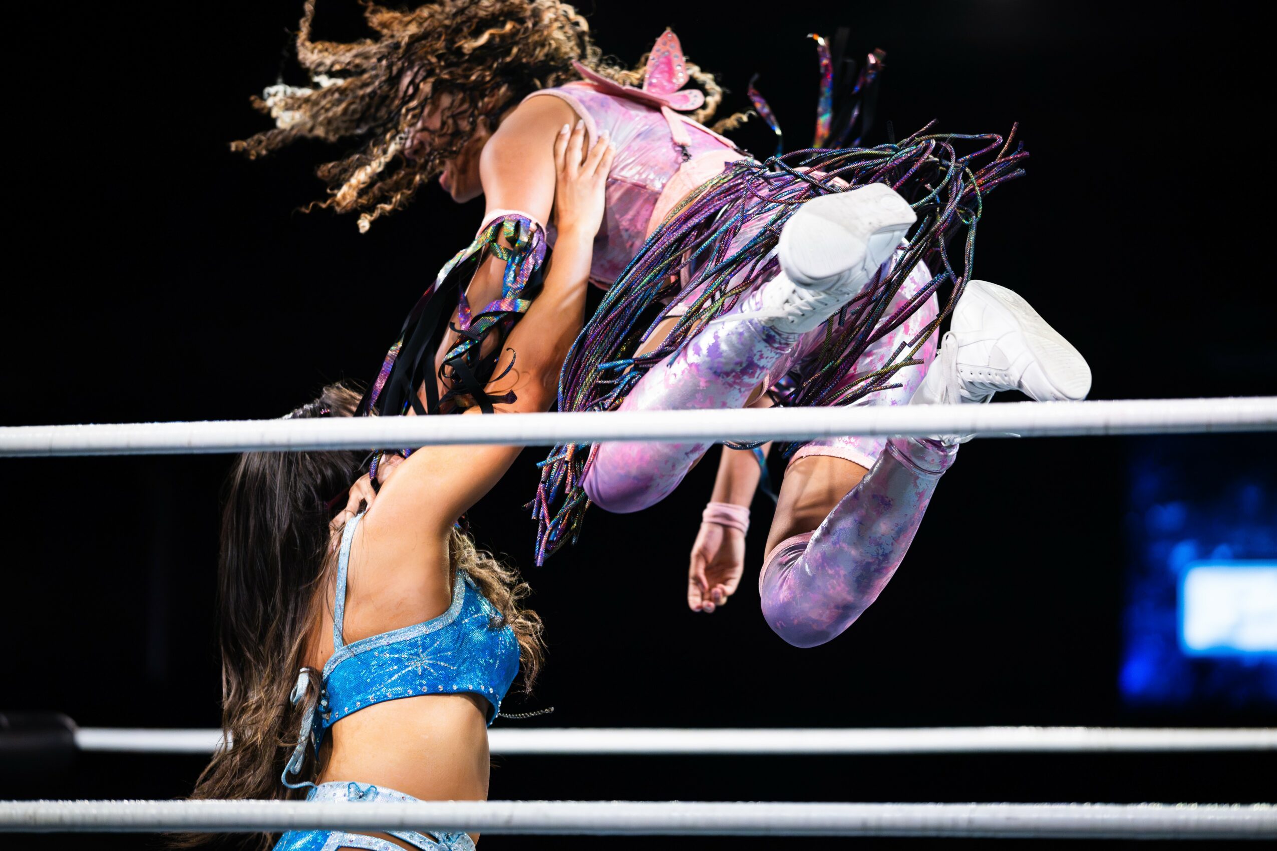 Two female wrestlers in a match, one in blue and the other in purple, mid-air in a wrestling ring.