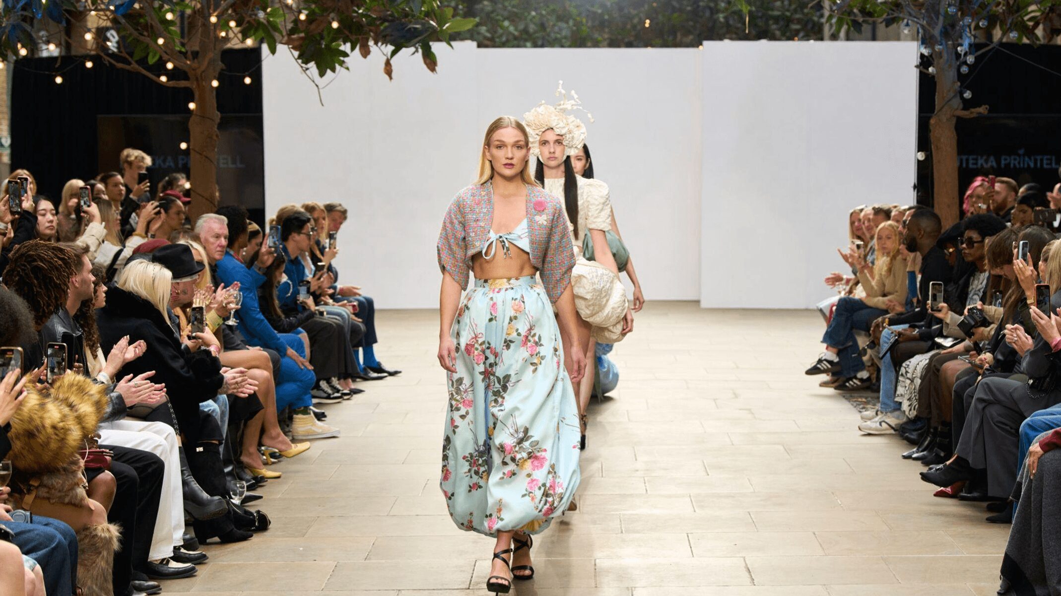 Fashion models walk down runway with audience on both sides, white backdrop behind them, outdoor setting, trees overhead.