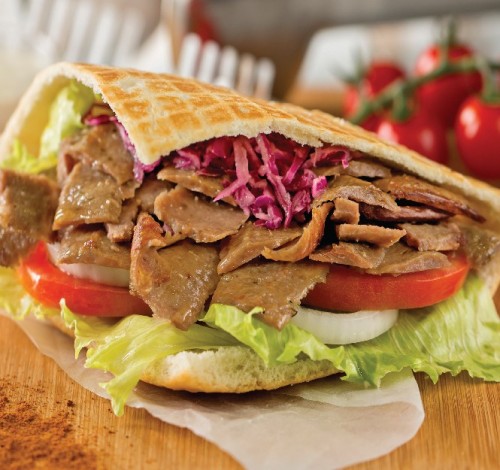 Sandwich with sliced meat, lettuce, tomato, and pickles on a toasted bread, with tomatoes in the background.