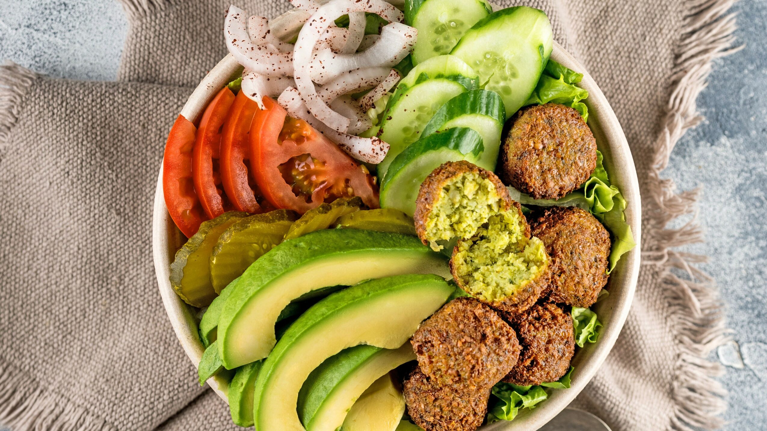 Bowl with sliced tomatoes, cucumbers, avocado, falafel, and pickles on a beige cloth background.