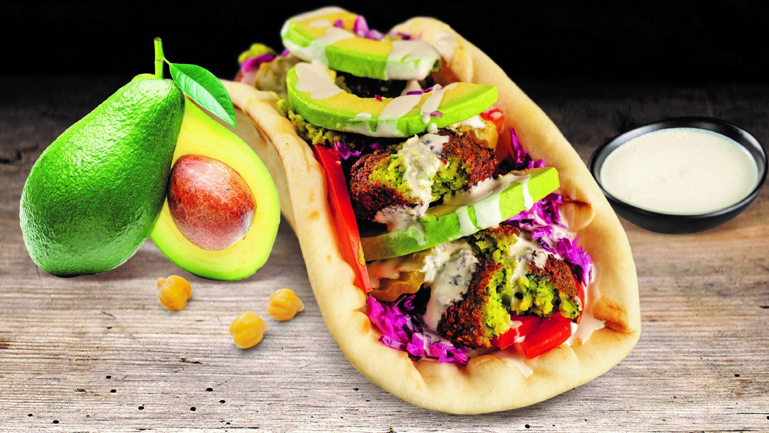 Tacos filled with vegetables and meat, with avocado and a small bowl of sauce on a wooden surface.