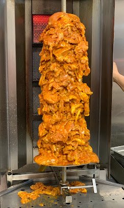 Vertical skewer of cooked, golden-brown meat rotating in a vertical rotisserie, with some meat slices falling off at the bottom.