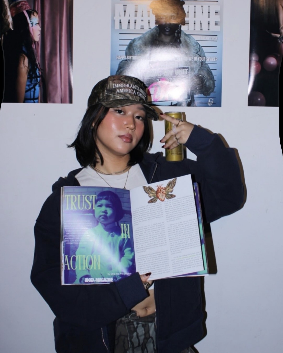 Young woman holding a magazine and a book, making a peace sign near her temple, with photos on the wall behind her.