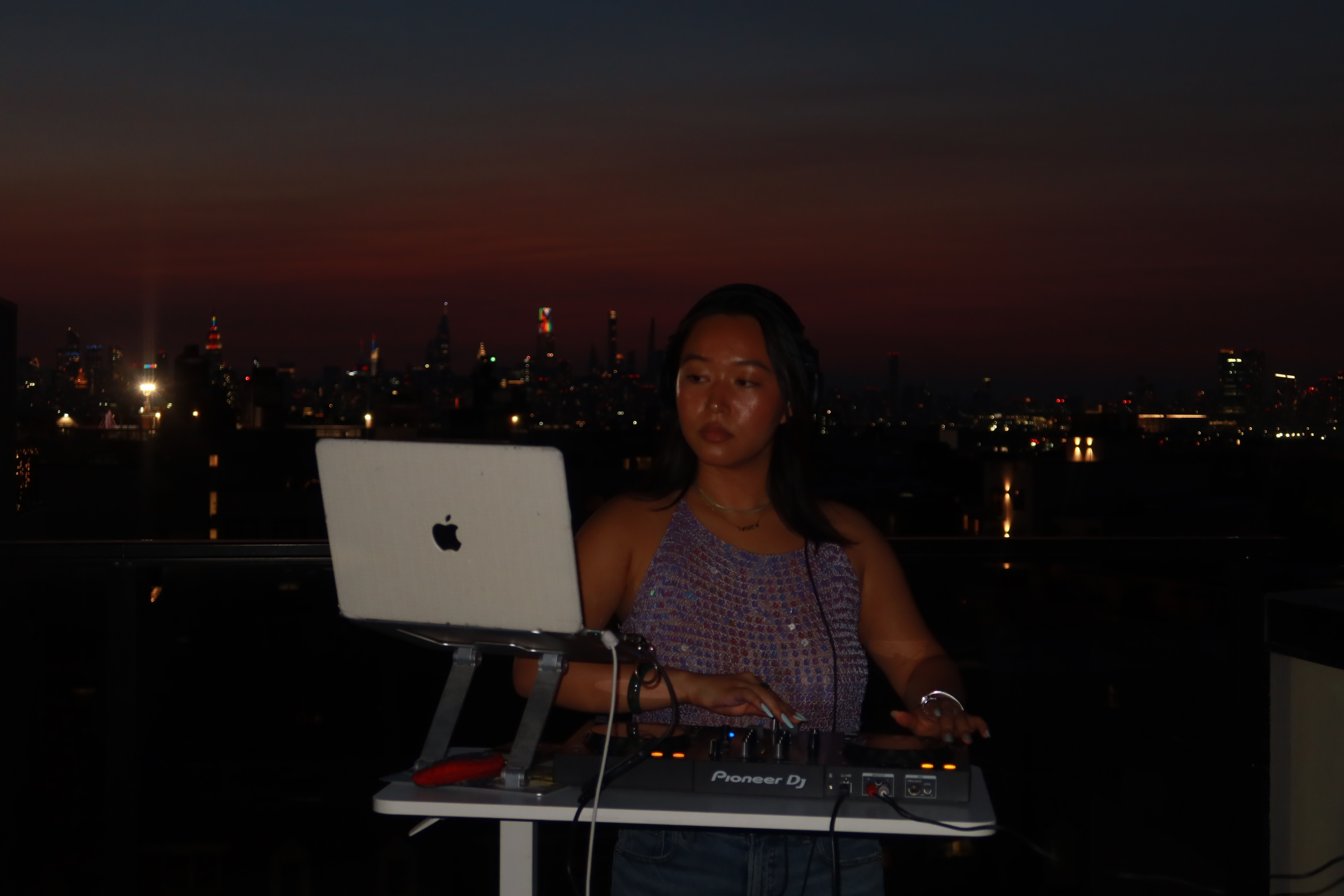 Person with long hair operating DJ equipment on rooftop at night with city skyline in background.