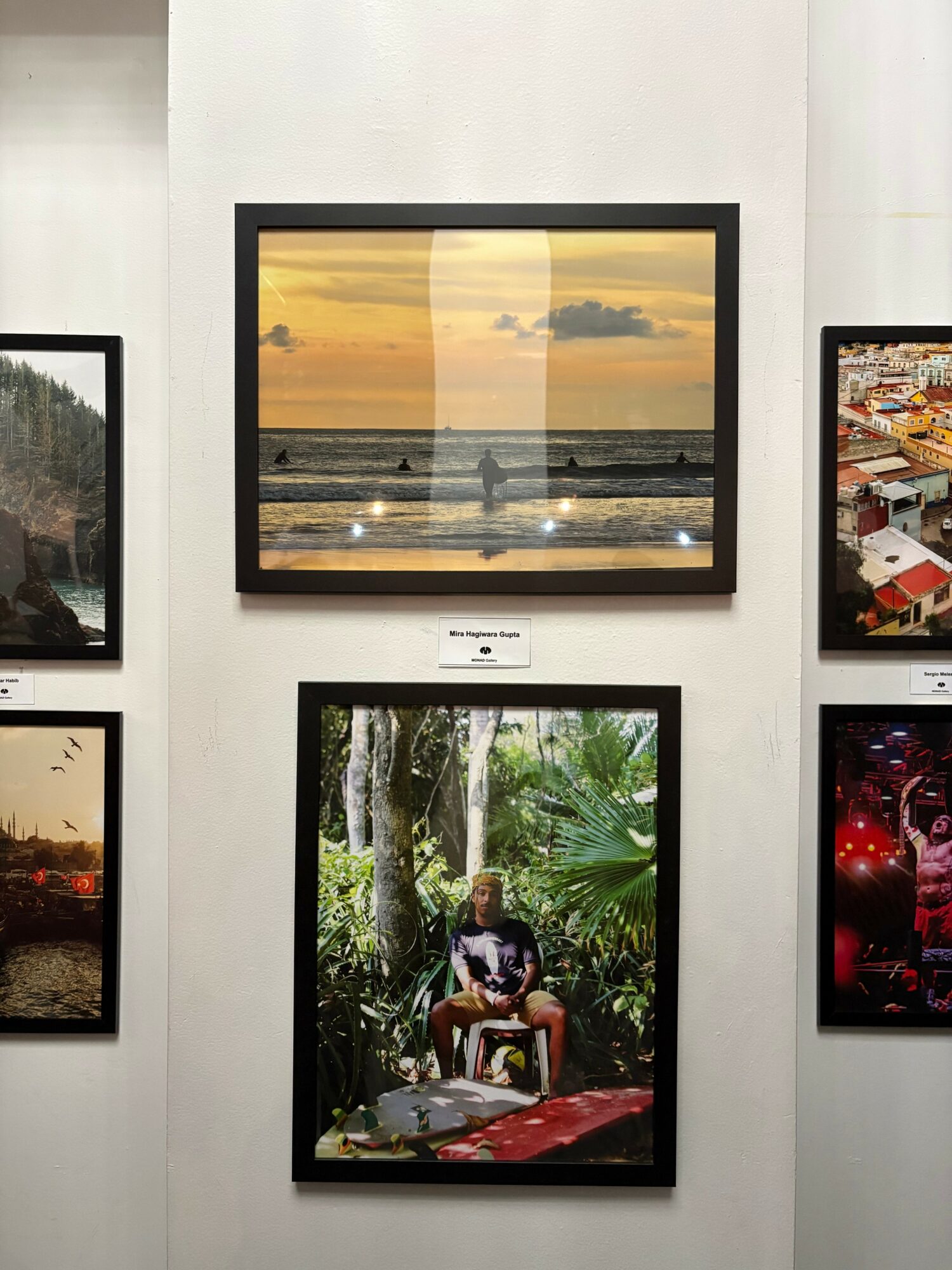 Framed sunset beach scene with clouds and ocean, person sitting in a lush green forest, and other framed images on white wall.