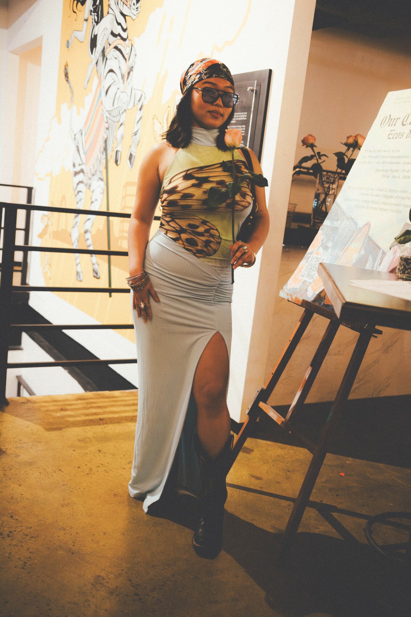 Woman in a colorful top and long white skirt with a slit, standing indoors near an art display.