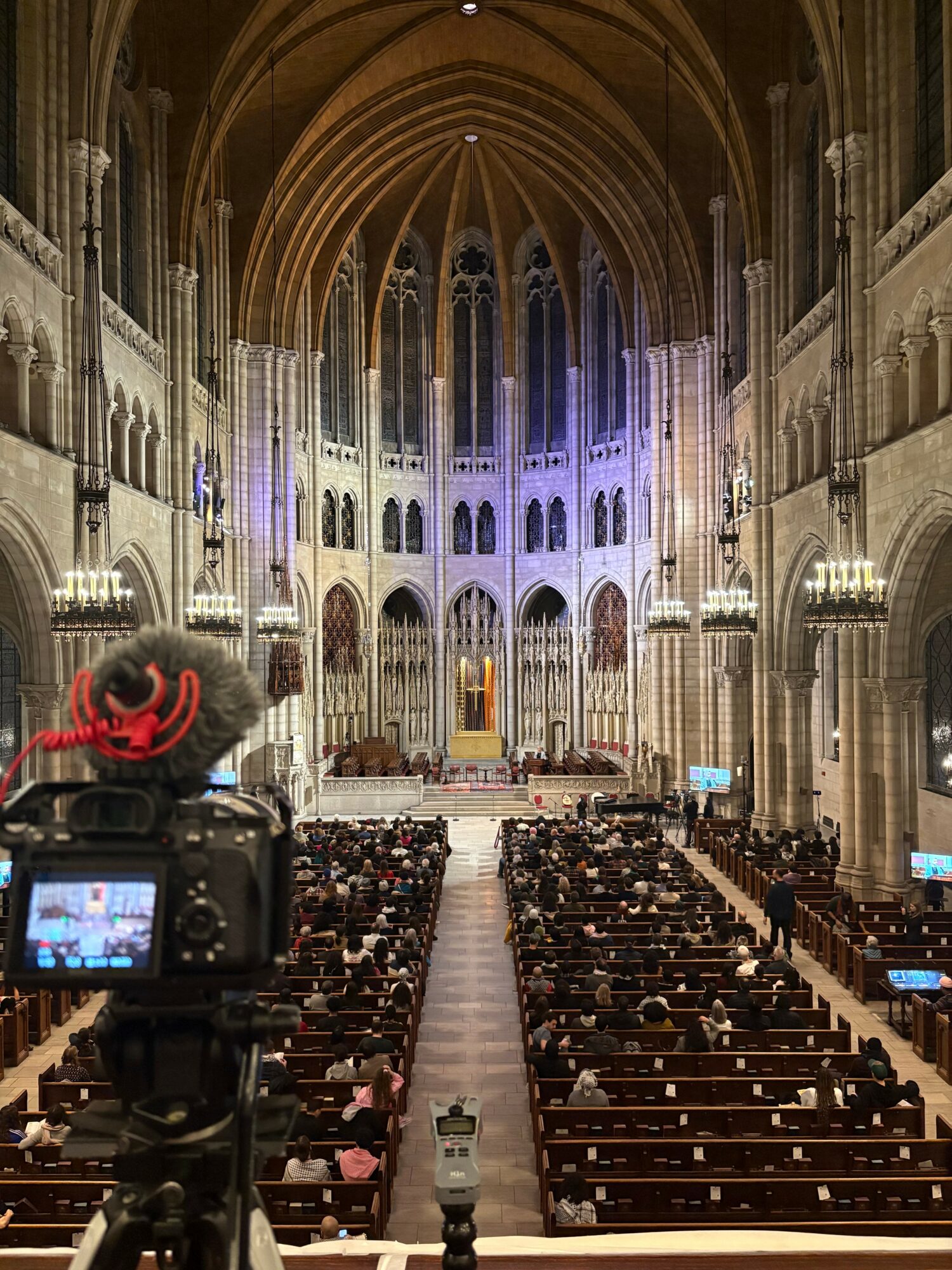 Interior of a large church or cathedral with high vaulted ceilings, ornate architecture, and an audience seated facing the altar.