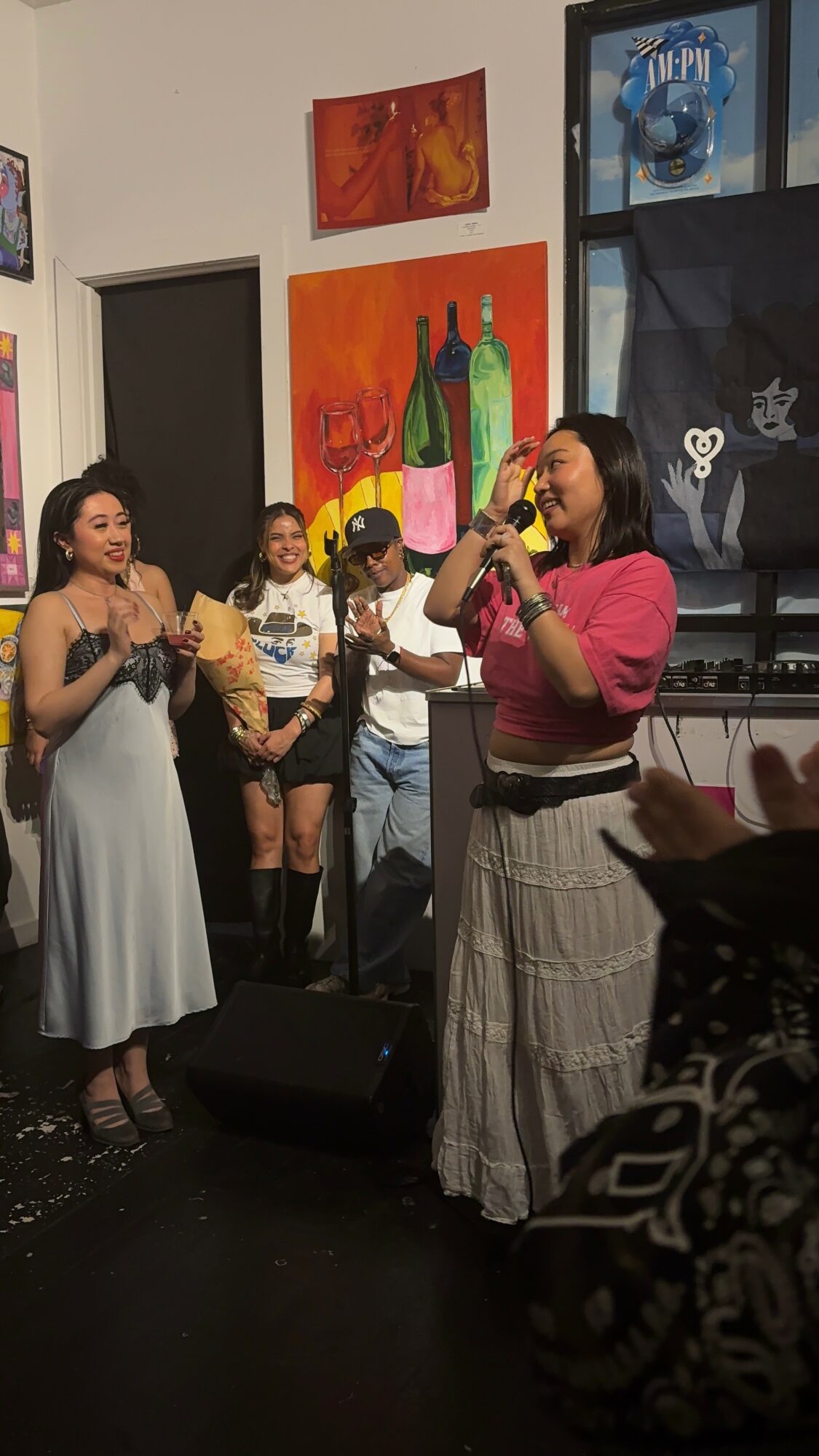 Two women speaking or singing on stage with a group of children in the background, colorful paintings on the wall.