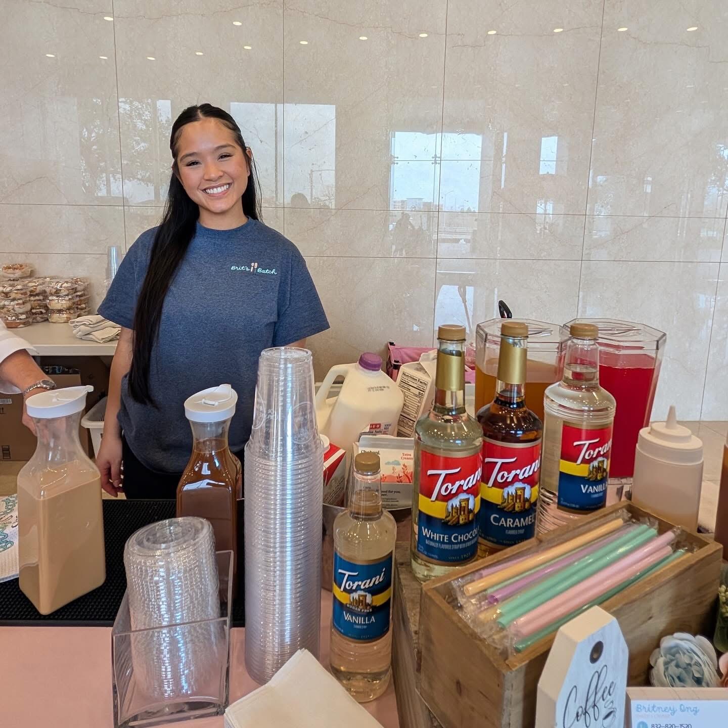 Smiling young woman behind a counter with bottles of Torani syrup and cups, in a bright indoor setting.