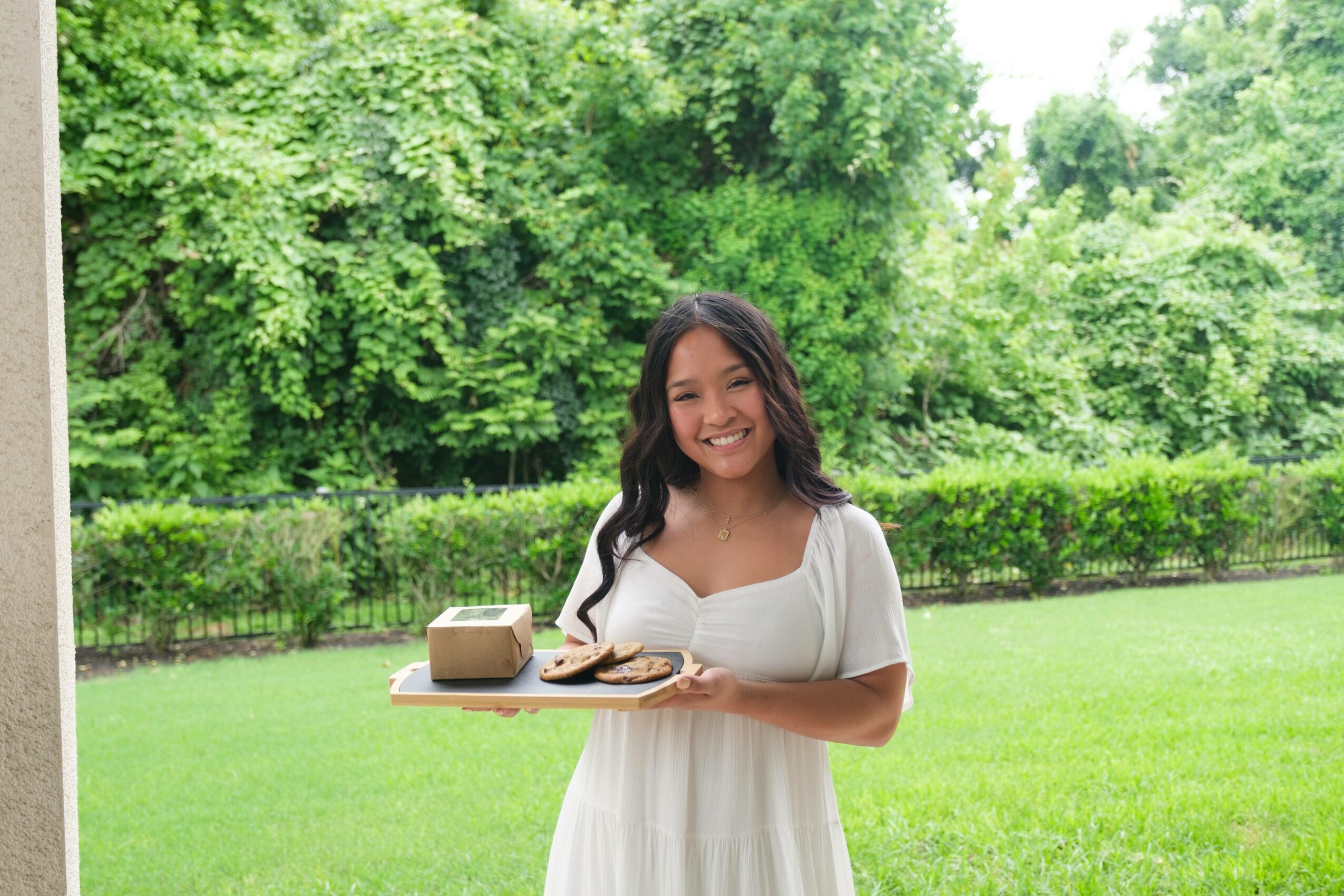 Smiling woman holding a tray with a cake outdoors in a green park.