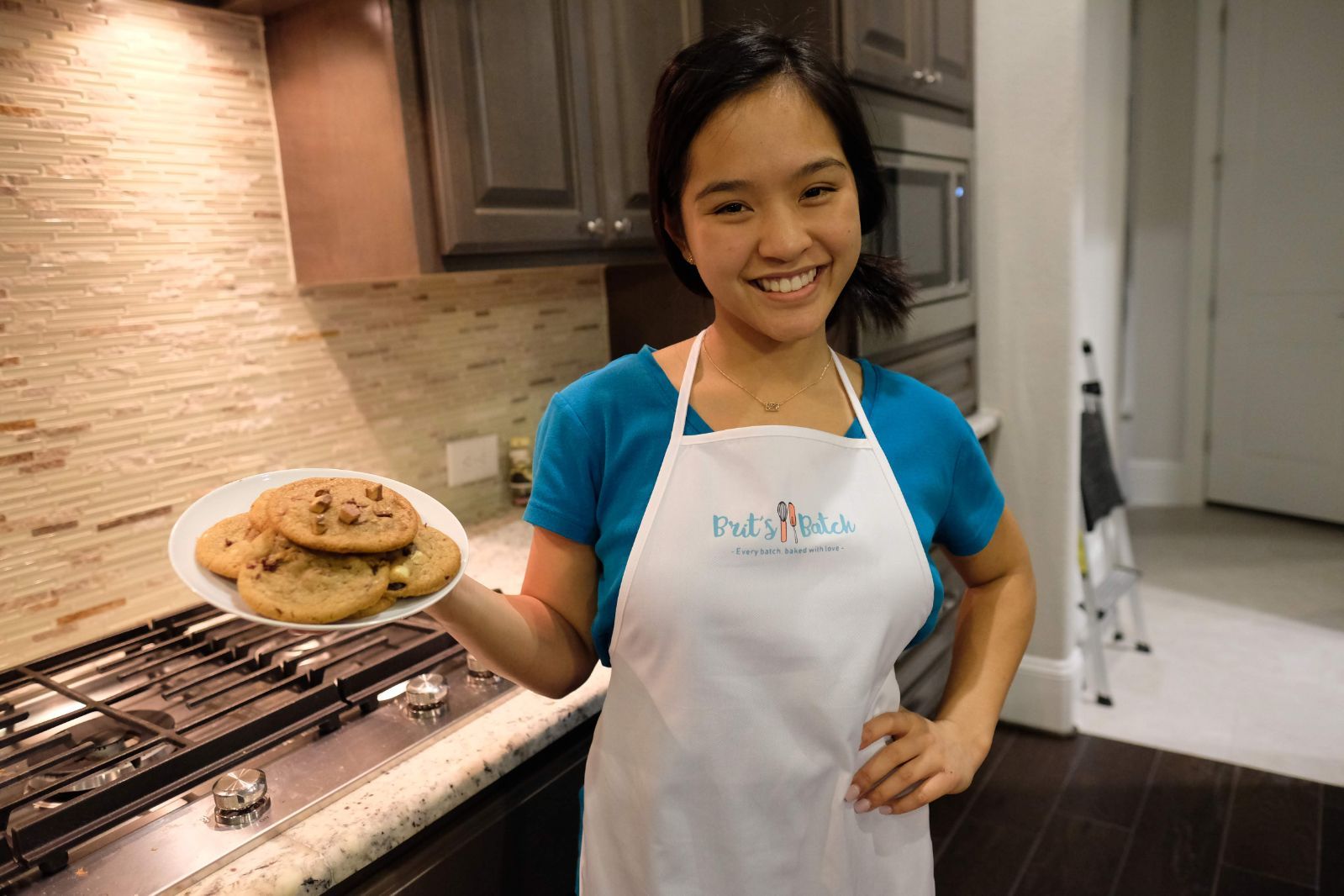 Young girl in a blue shirt and apron holding a plate of cookies in a kitchen, smiling.