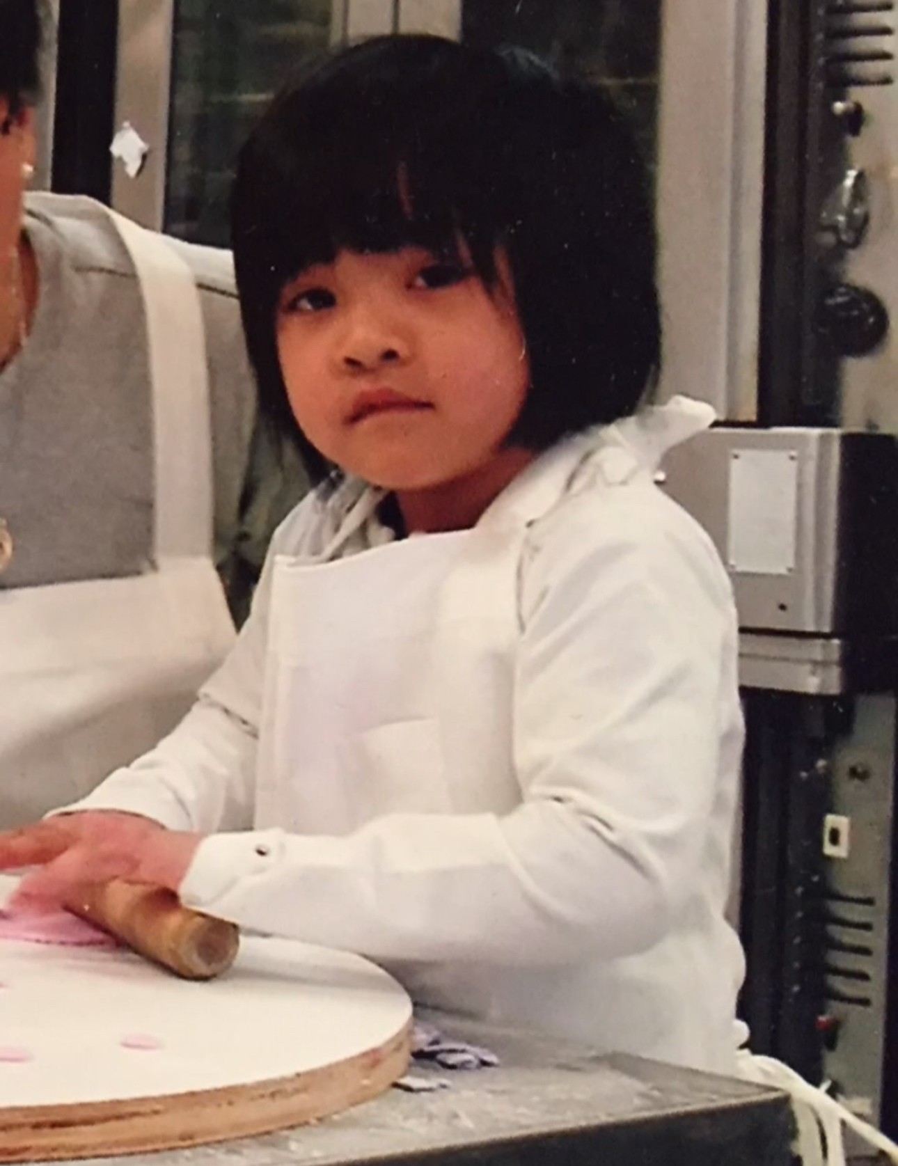 Child with black hair and white shirt, sitting at a table with a round object, in a room with equipment in the background.