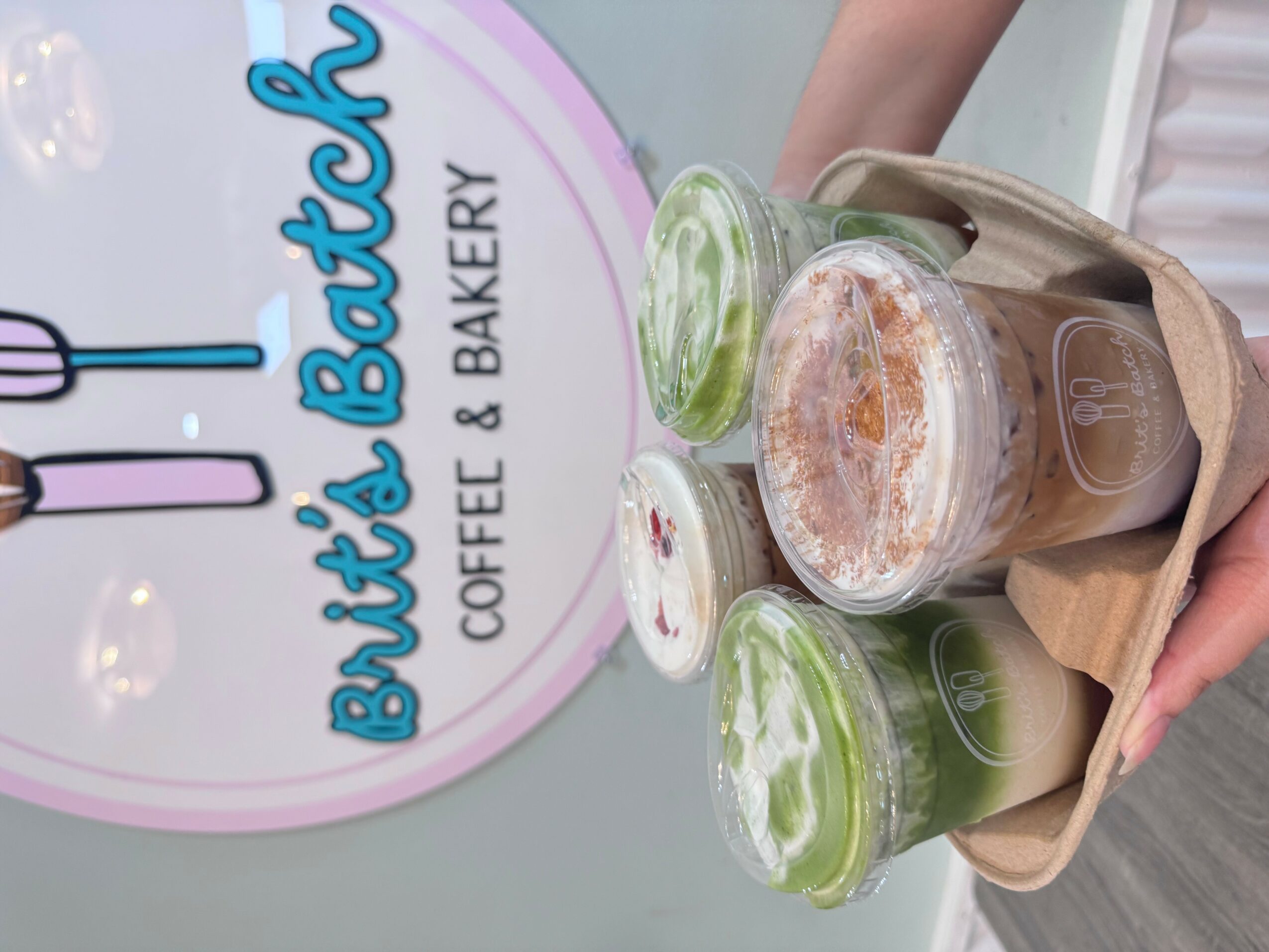 Tray with three green drinks and a brown beverage, in front of a bakery sign, held by a person.