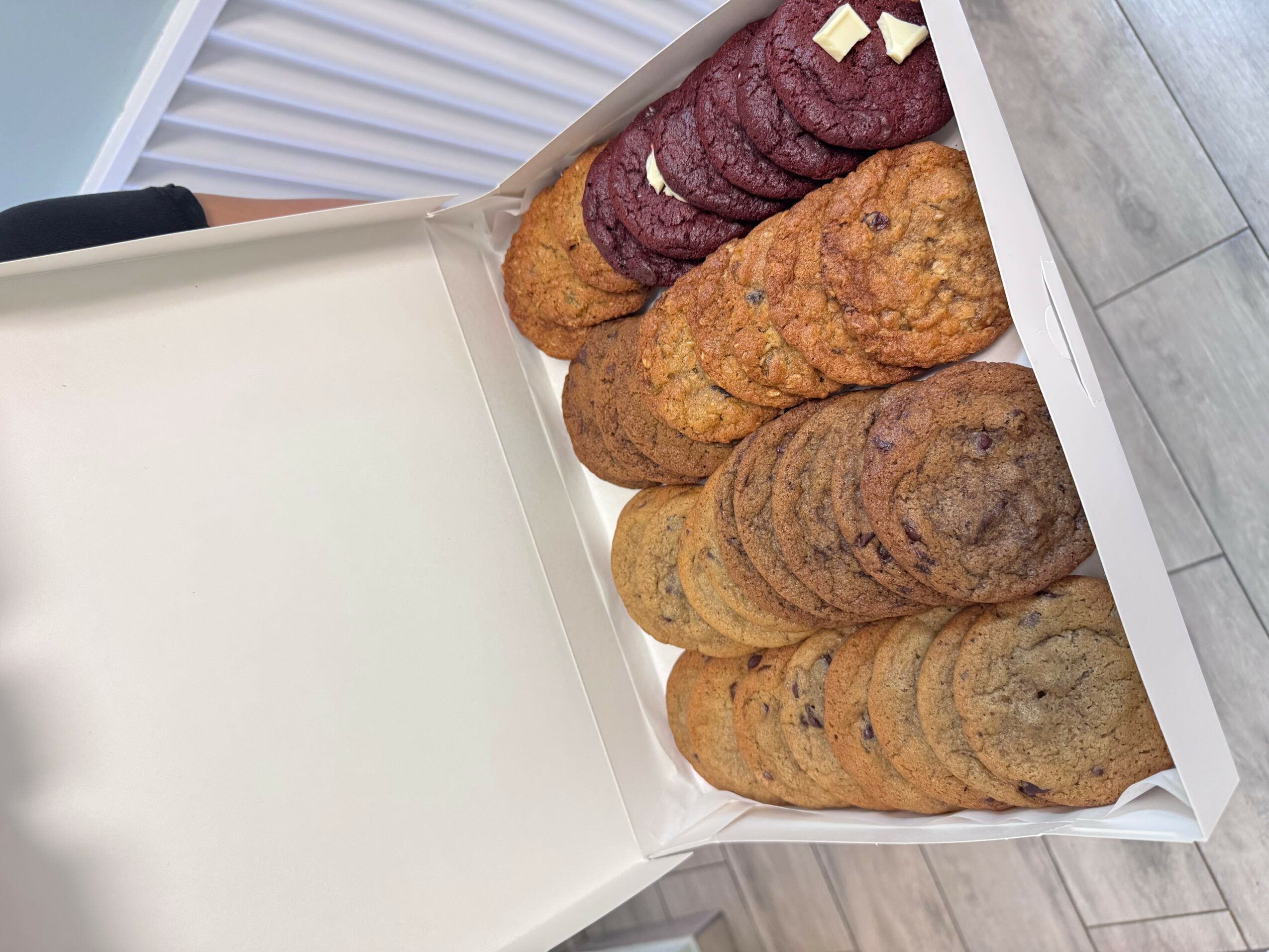 Box of assorted cookies, including chocolate chip, oatmeal, and peanut butter, on a wooden floor.