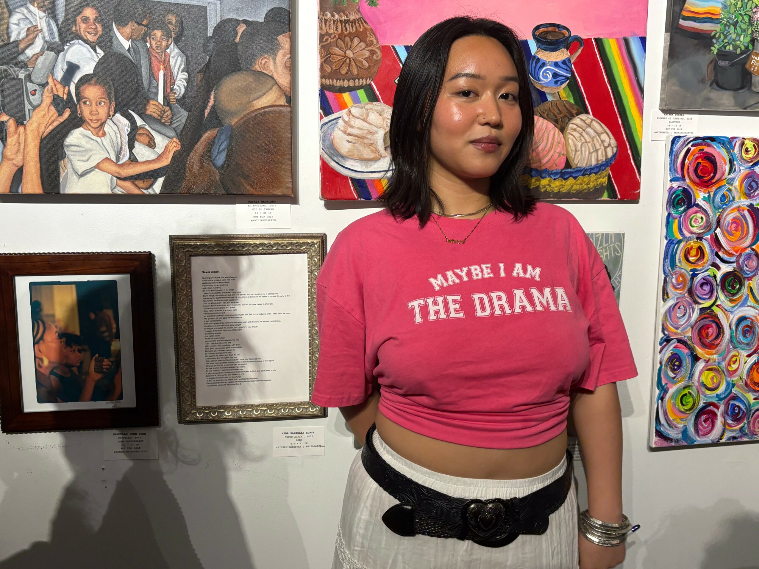 Young woman with shoulder-length black hair wearing a pink crop top with white text, standing in front of colorful artwork.