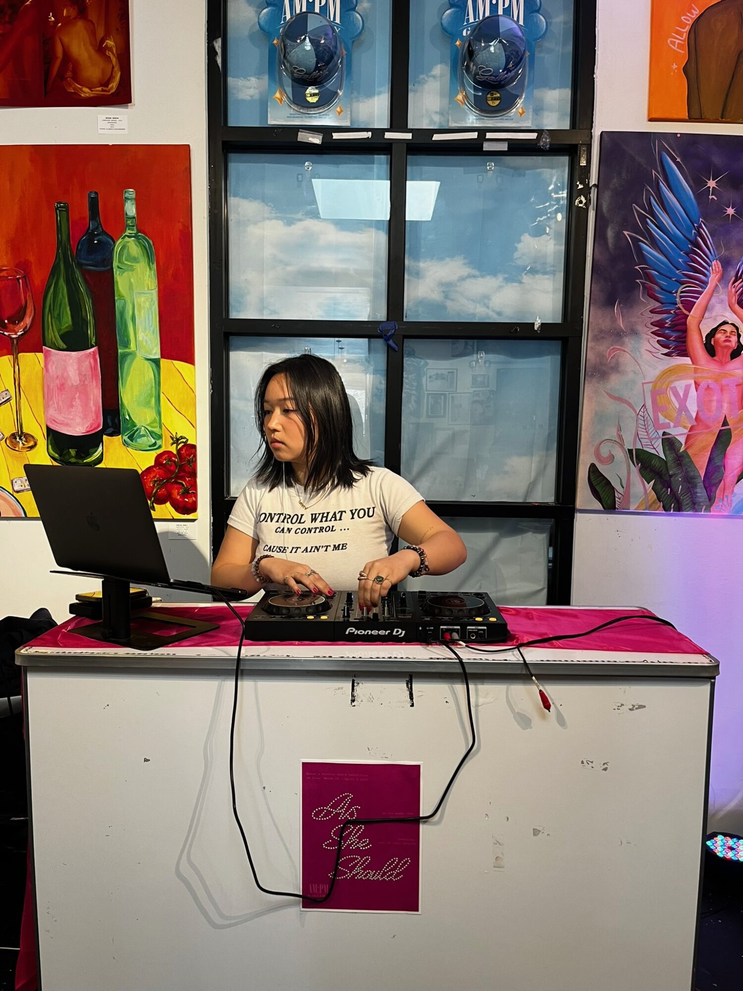 Young woman DJing at a table with a laptop and equipment, colorful paintings on the wall behind her.