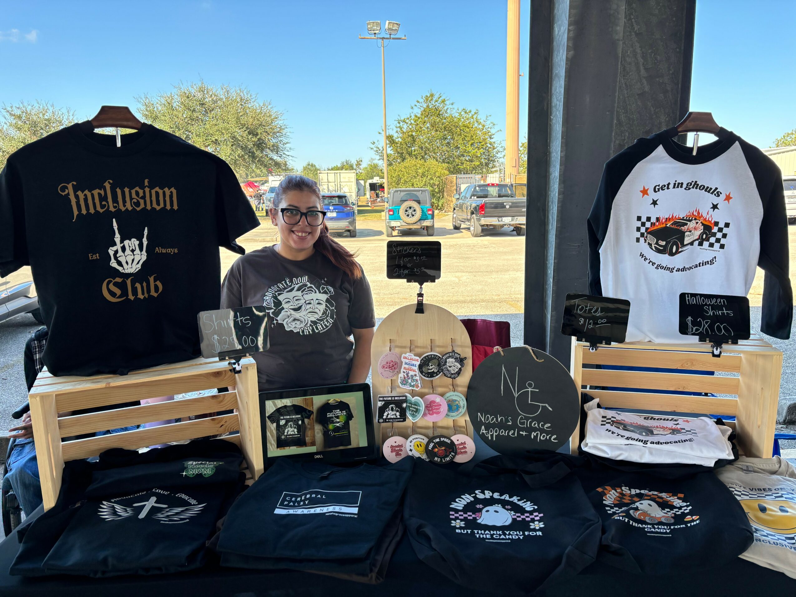 Woman standing behind a table with black and white shirts, promotional items, and signs at an outdoor event.