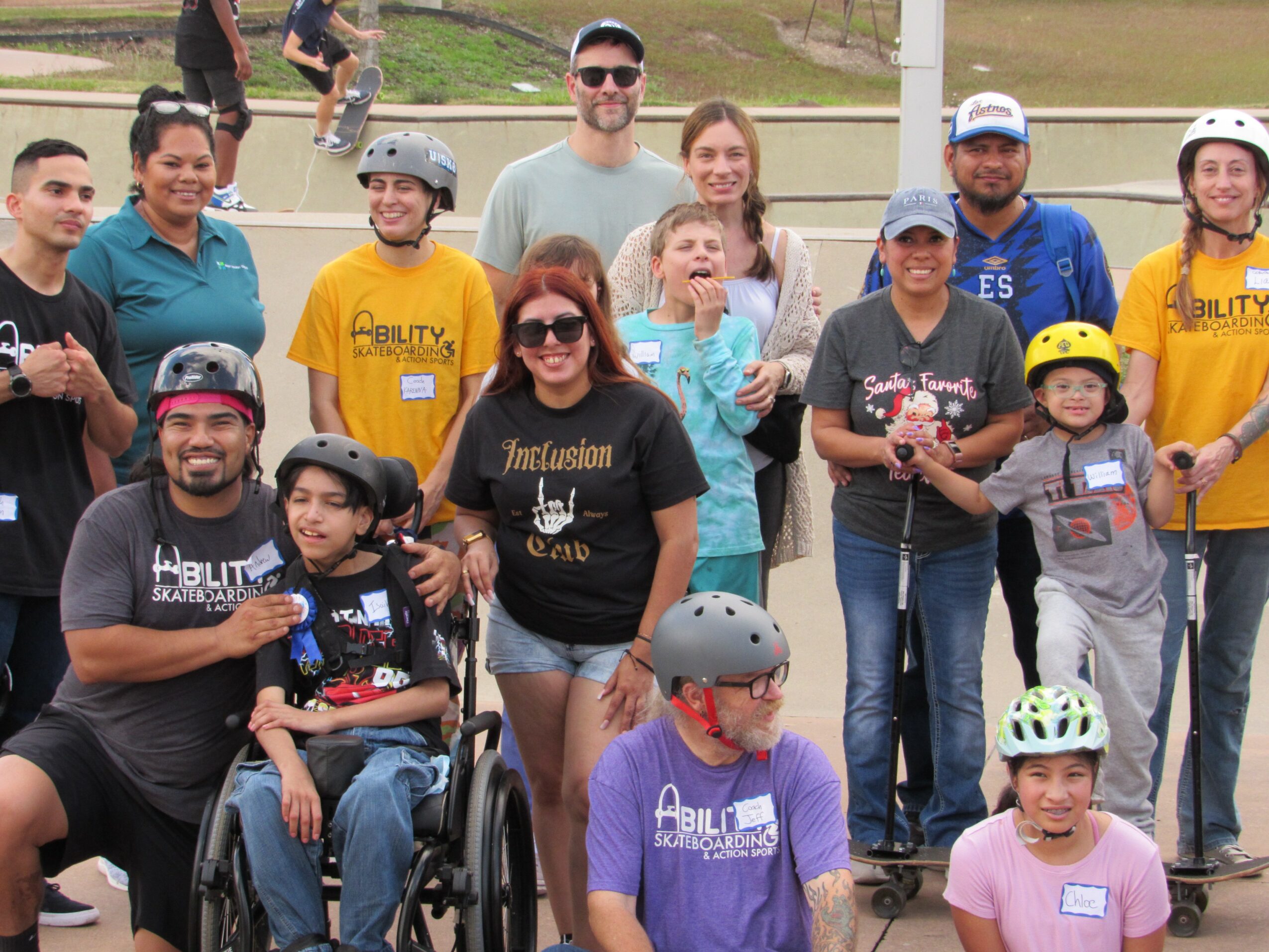 Group of people, some in wheelchairs, wearing helmets and casual clothing, outdoors on a grassy area, smiling for a photo.