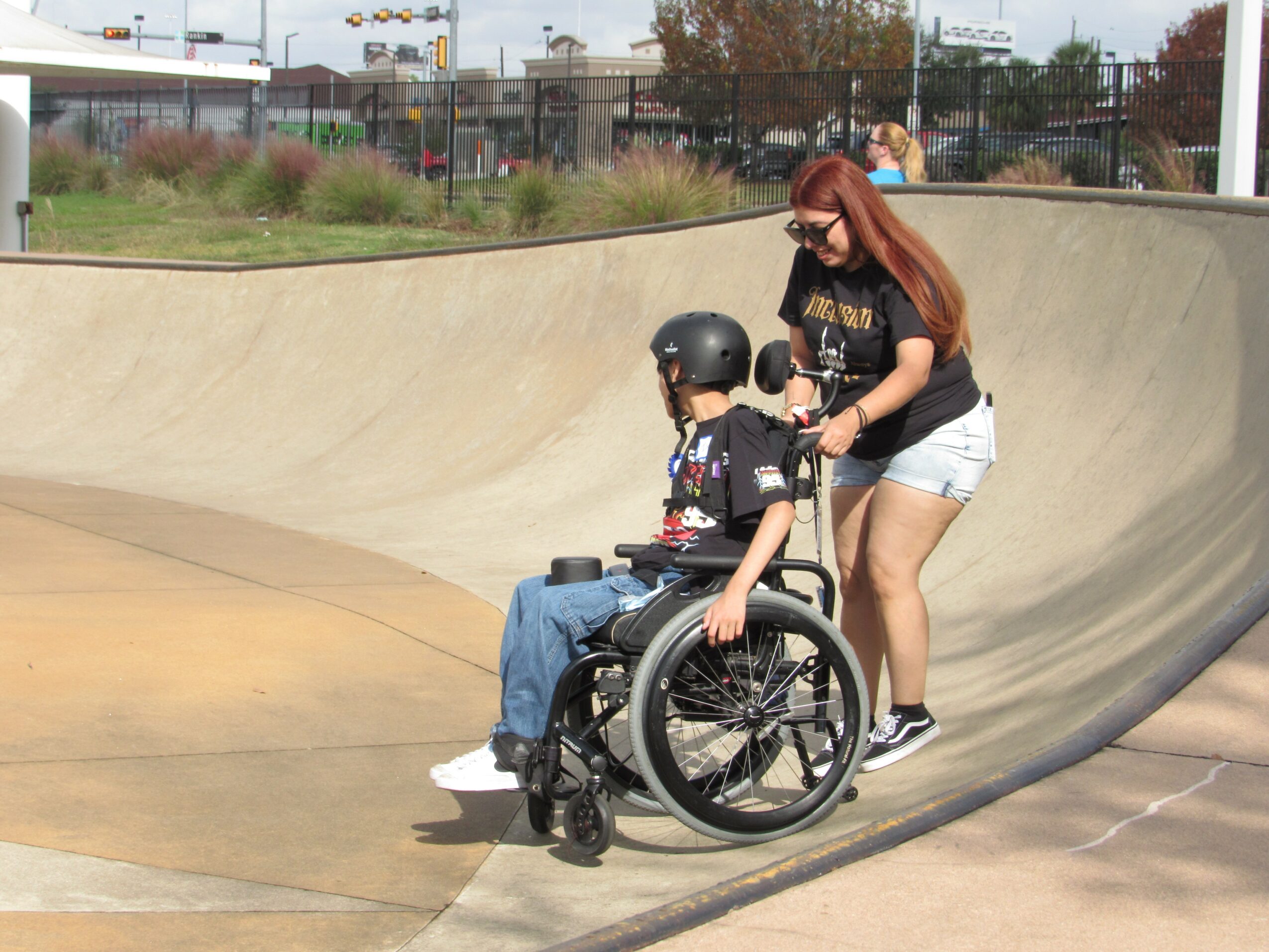 A woman with long hair and sunglasses helps a person in a wheelchair at a skate park, with a concrete bowl and outdoor setting.