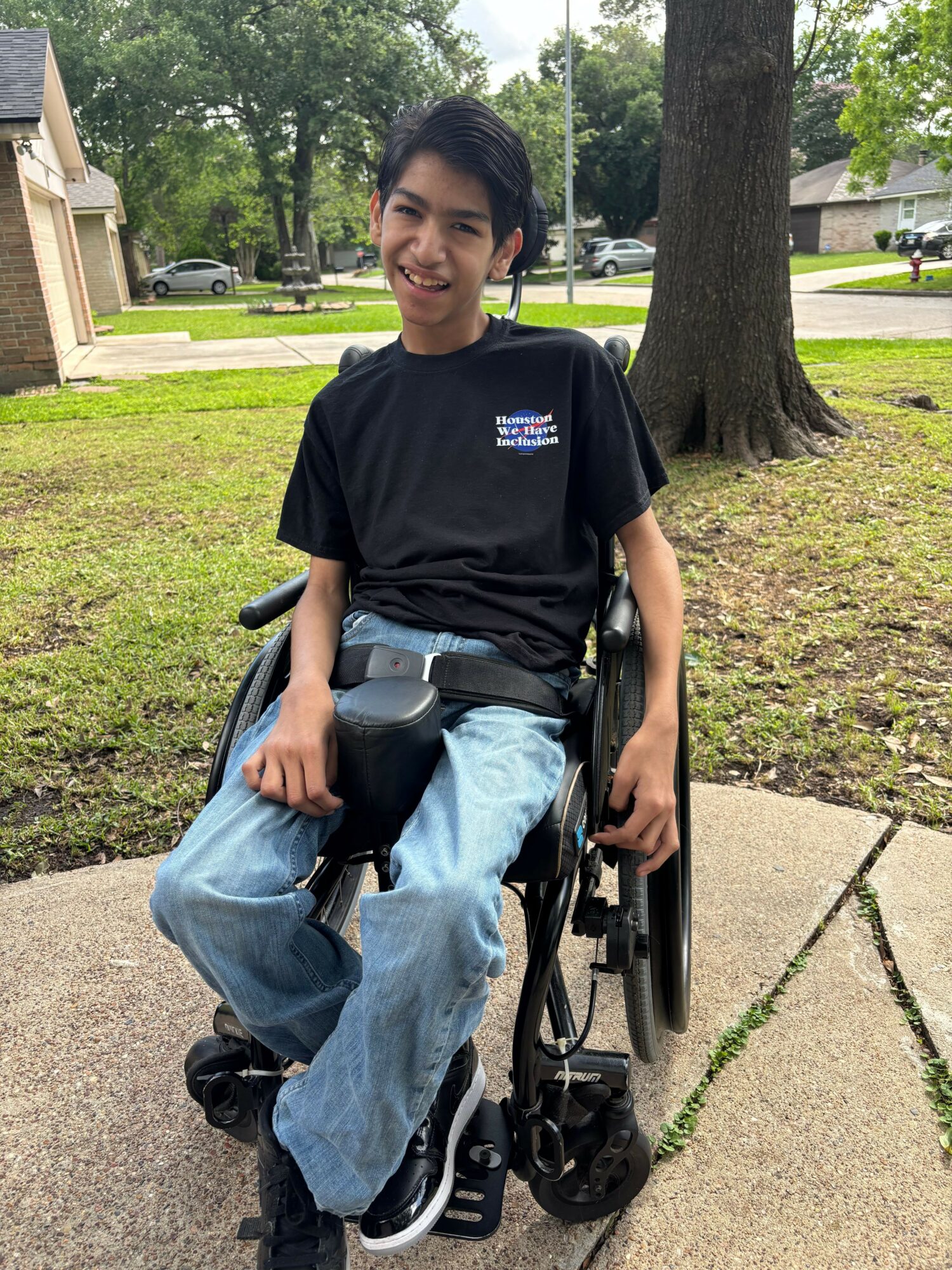 Young woman in wheelchair on sidewalk, smiling, wearing black shirt and jeans, in park with trees and grass.