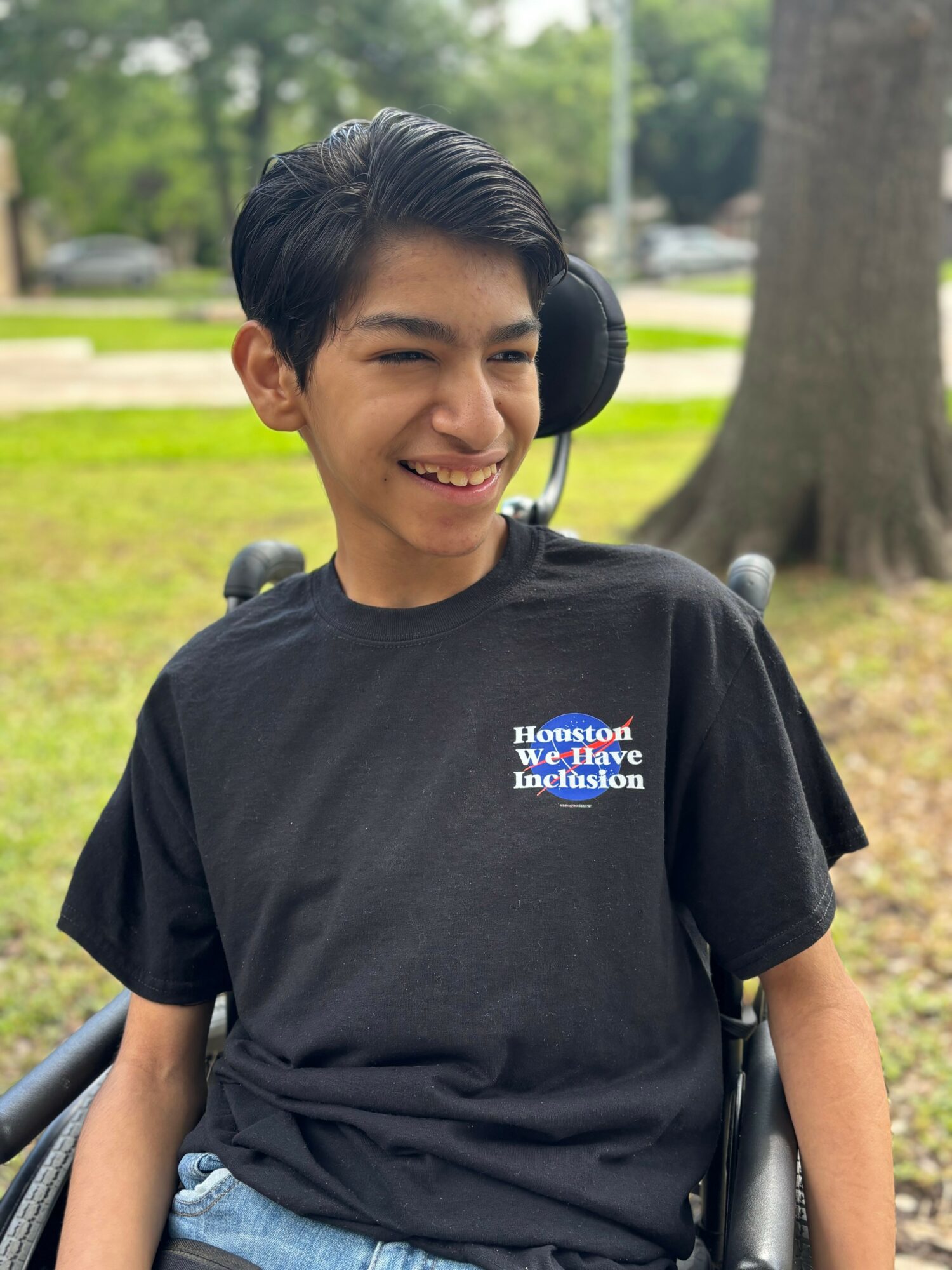 Young person smiling outdoors, wearing a black T-shirt with text, sitting in a wheelchair, trees and grass in background.