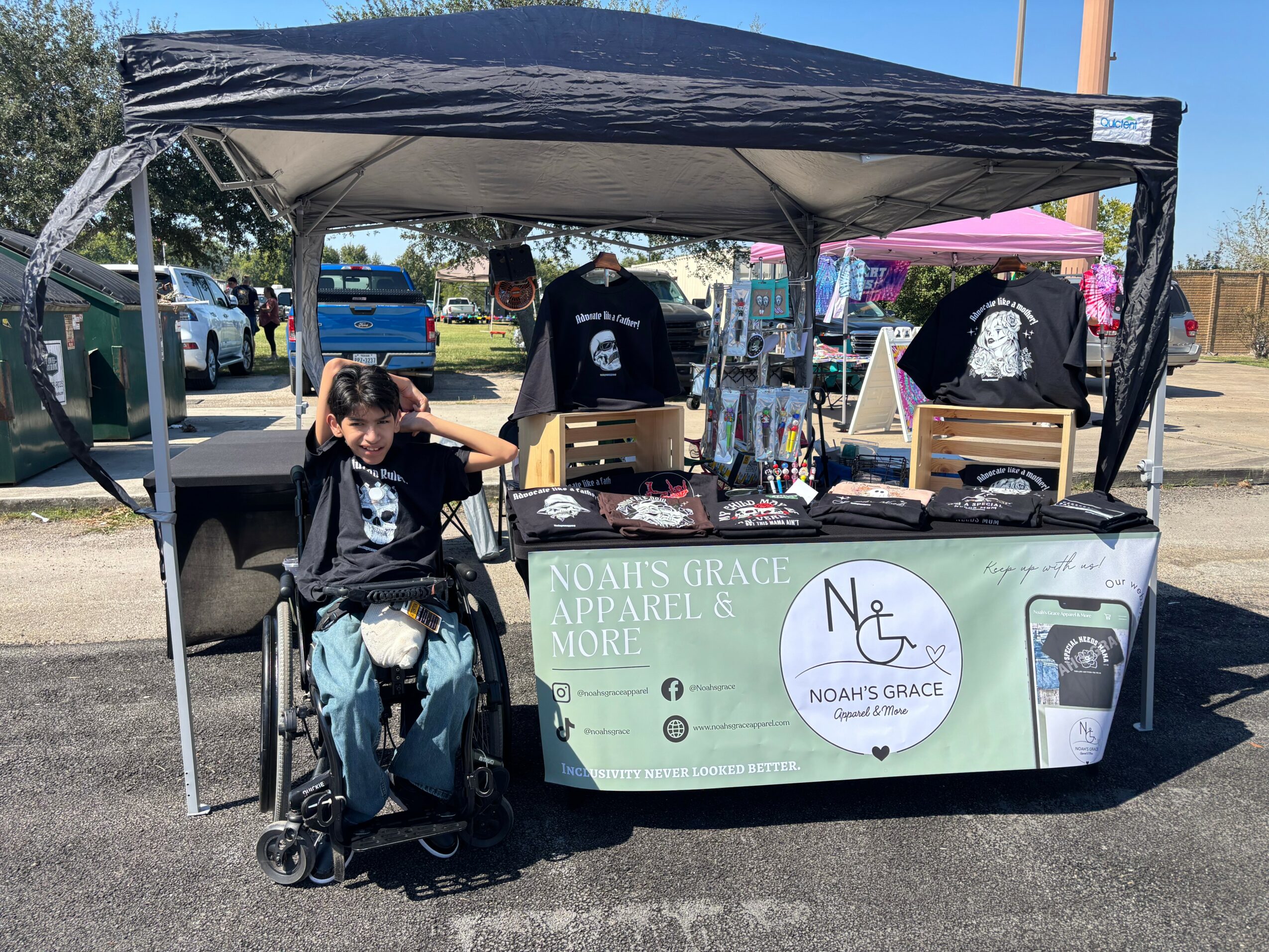 Person sitting in a wheelchair in front of a booth with a canopy at an outdoor event, with people and cars in background.