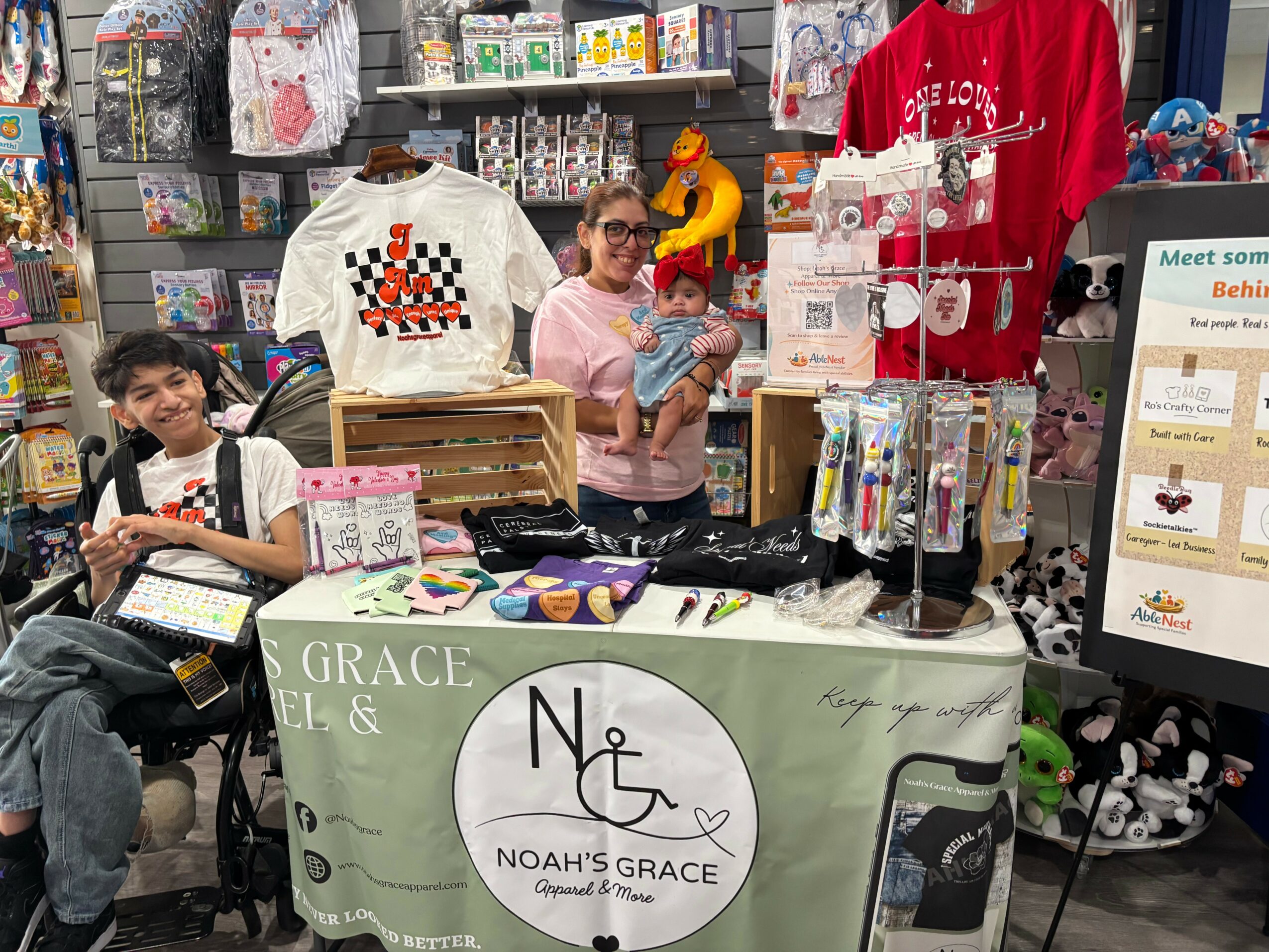 Two women and a child at a store counter with clothing and toys, smiling and interacting, surrounded by shelves and displays.