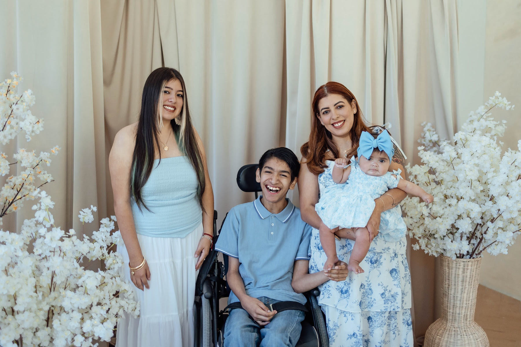 Four people, including a young girl in a wheelchair, pose in front of white curtains with white floral arrangements, smiling.