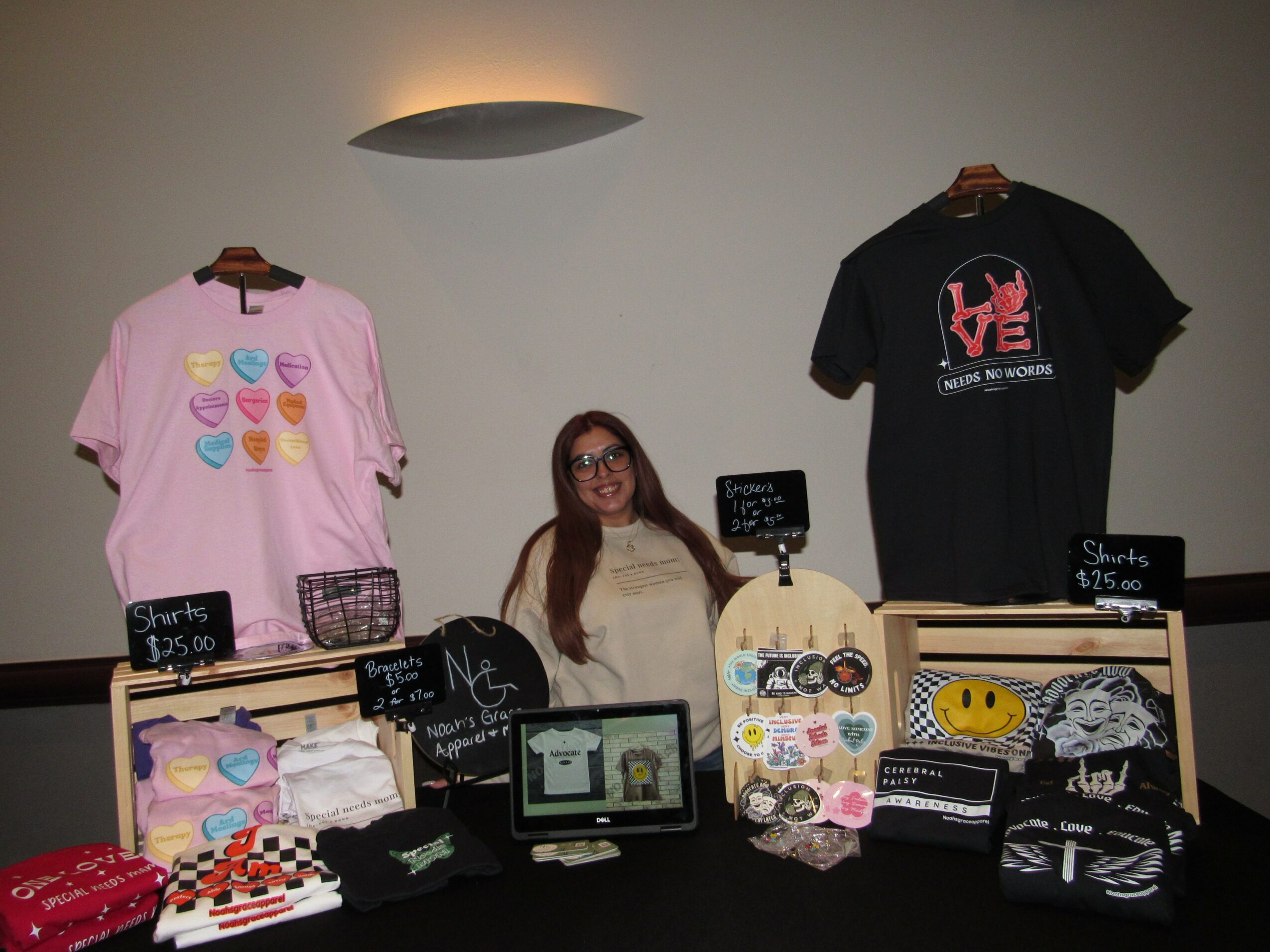 Woman sitting behind a table with colorful merchandise, T-shirts hanging on wall, and small signs, in a shop or market setting.