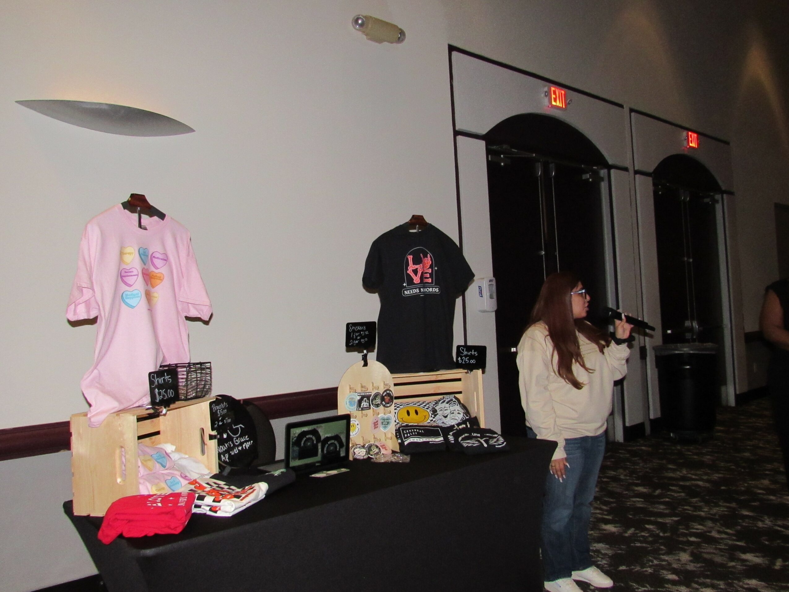 Person taking a photo of merchandise table with clothing and accessories, pink and black shirts displayed on wall, indoor setting.