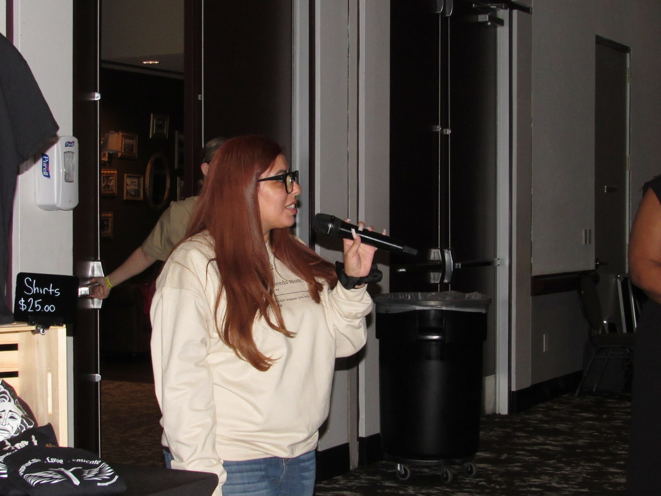 Woman with long red hair and glasses holding a microphone, standing indoors near a black trash bin.