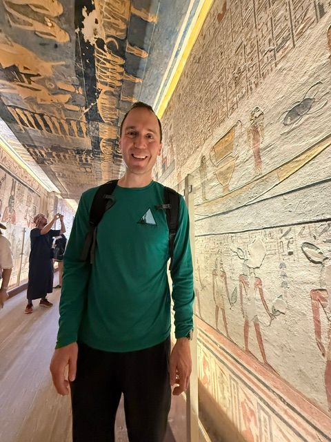 Person standing in an art gallery with ancient wall paintings, wearing a green jacket and black pants.
