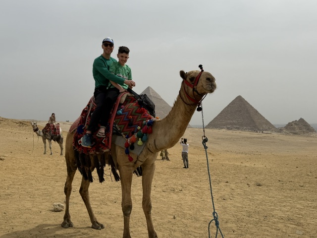 Two people riding a camel in a desert with pyramids in the background, one person wearing sunglasses and a green shirt.