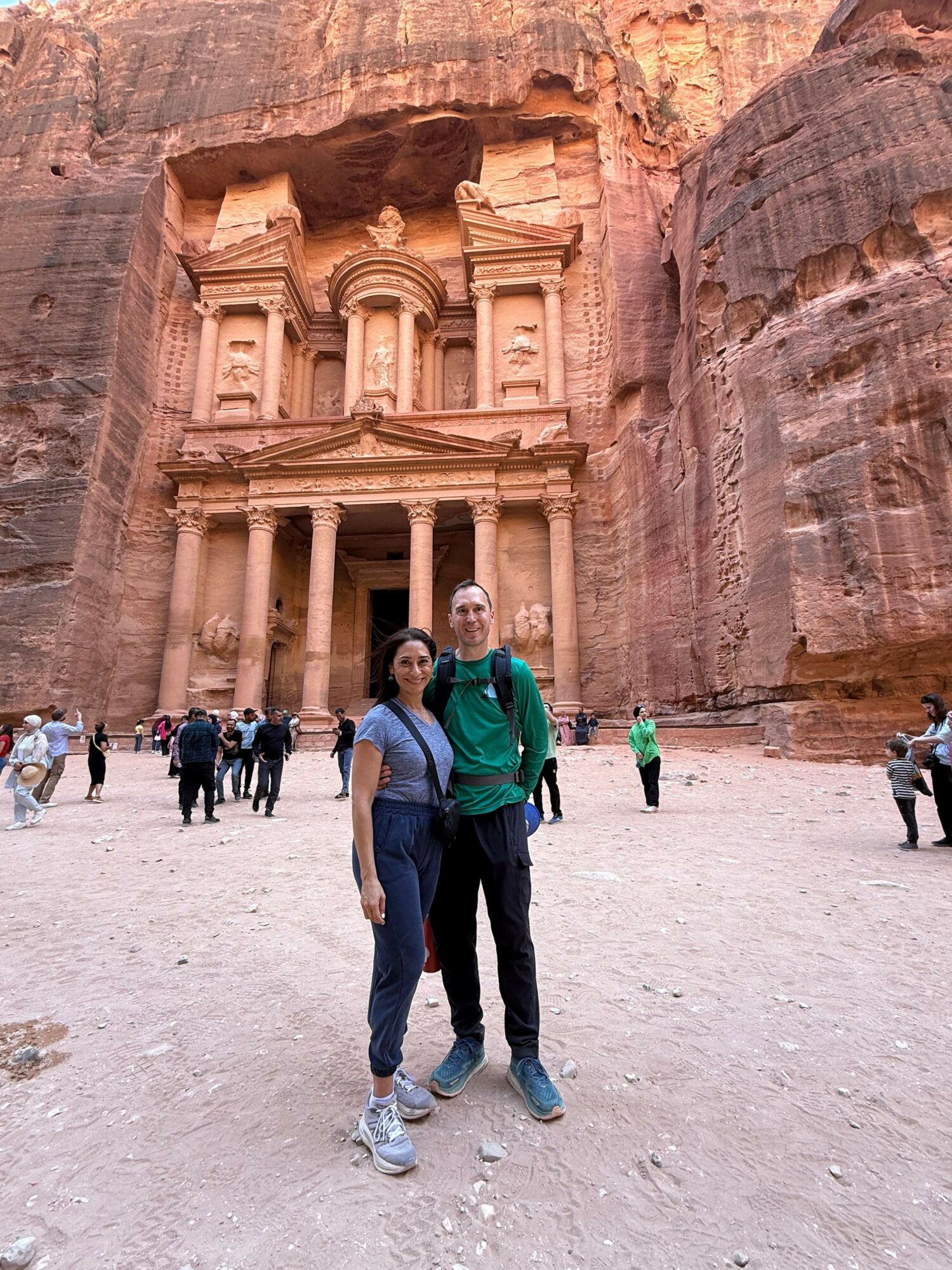 Two people walk in front of a carved rock face with an ancient structure, surrounded by a desert landscape.