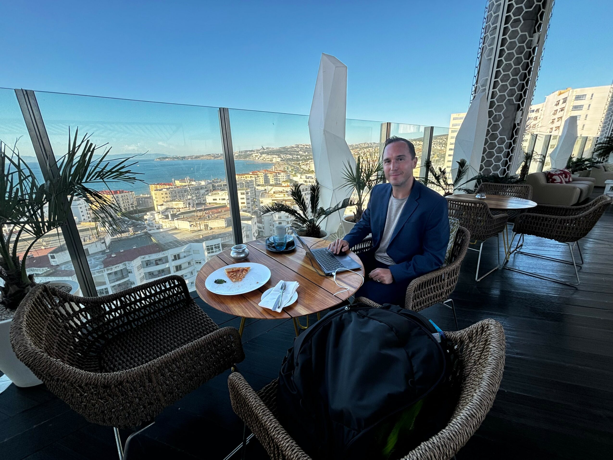 Man in a suit sitting at a table on a balcony with city view, plants, and a glass railing, holding a tablet.
