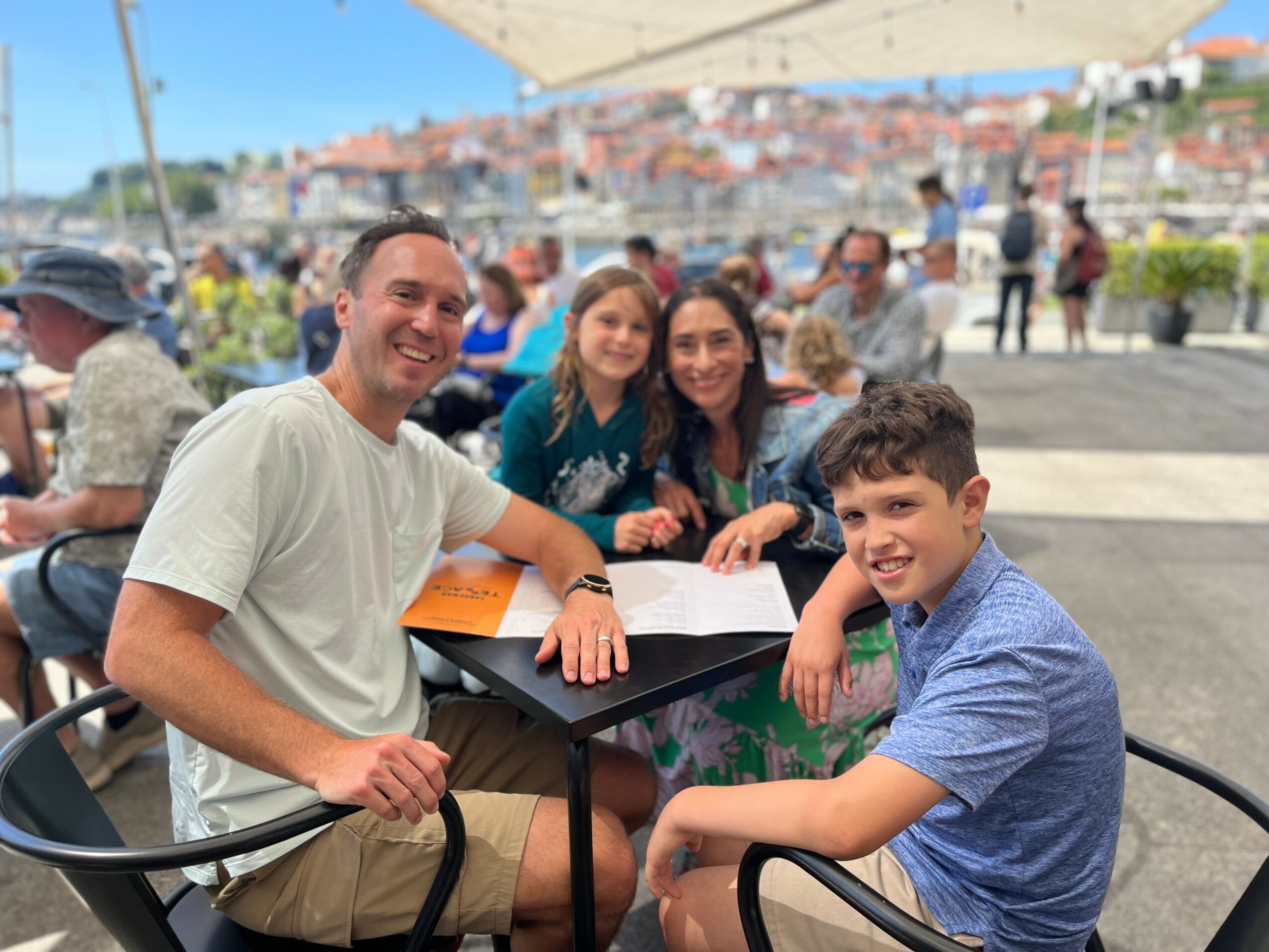 Four people sitting at a table outdoors, smiling, with a cityscape in the background.