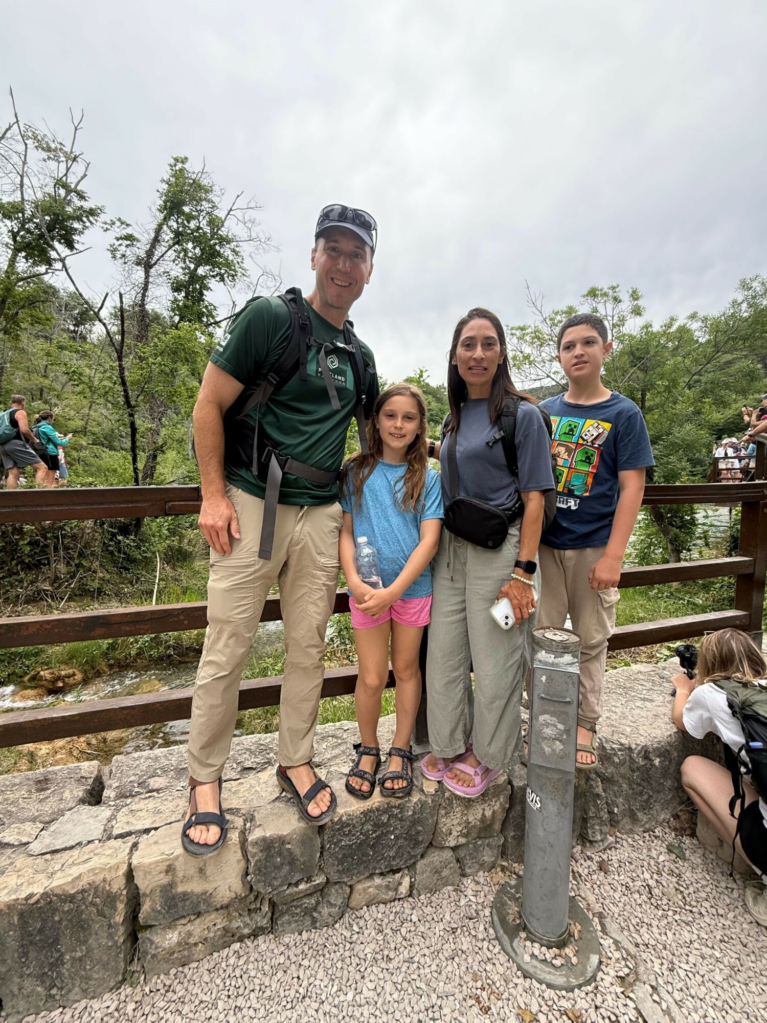 Four people standing outdoors on a cobblestone path with trees and a wooden railing in the background.