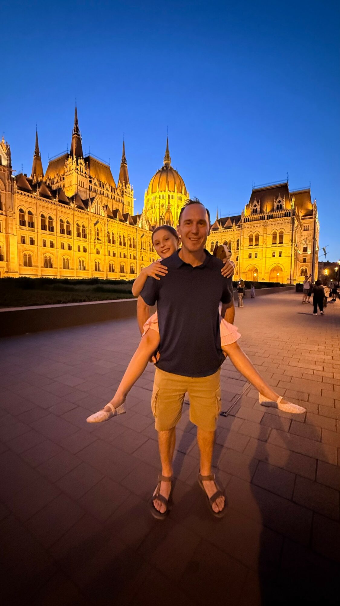 Two people standing outdoors in front of illuminated historic buildings at dusk, smiling and embracing.