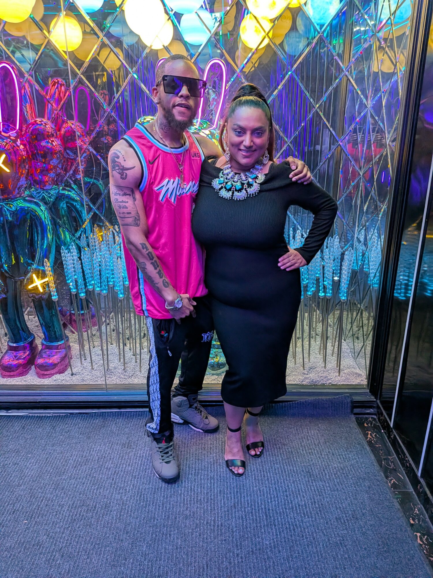 Two people stand in front of colorful neon lights and decorations, smiling and posing for the photo.