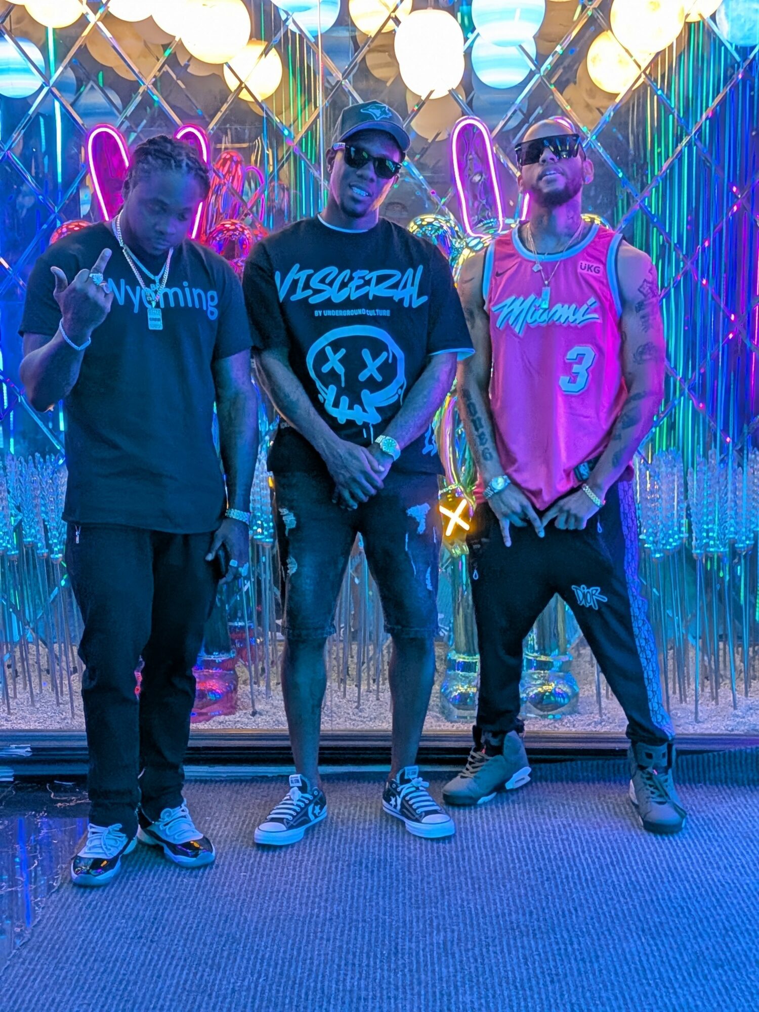 Three young men stand in front of a colorful, illuminated background with hanging lights and reflective decorations.