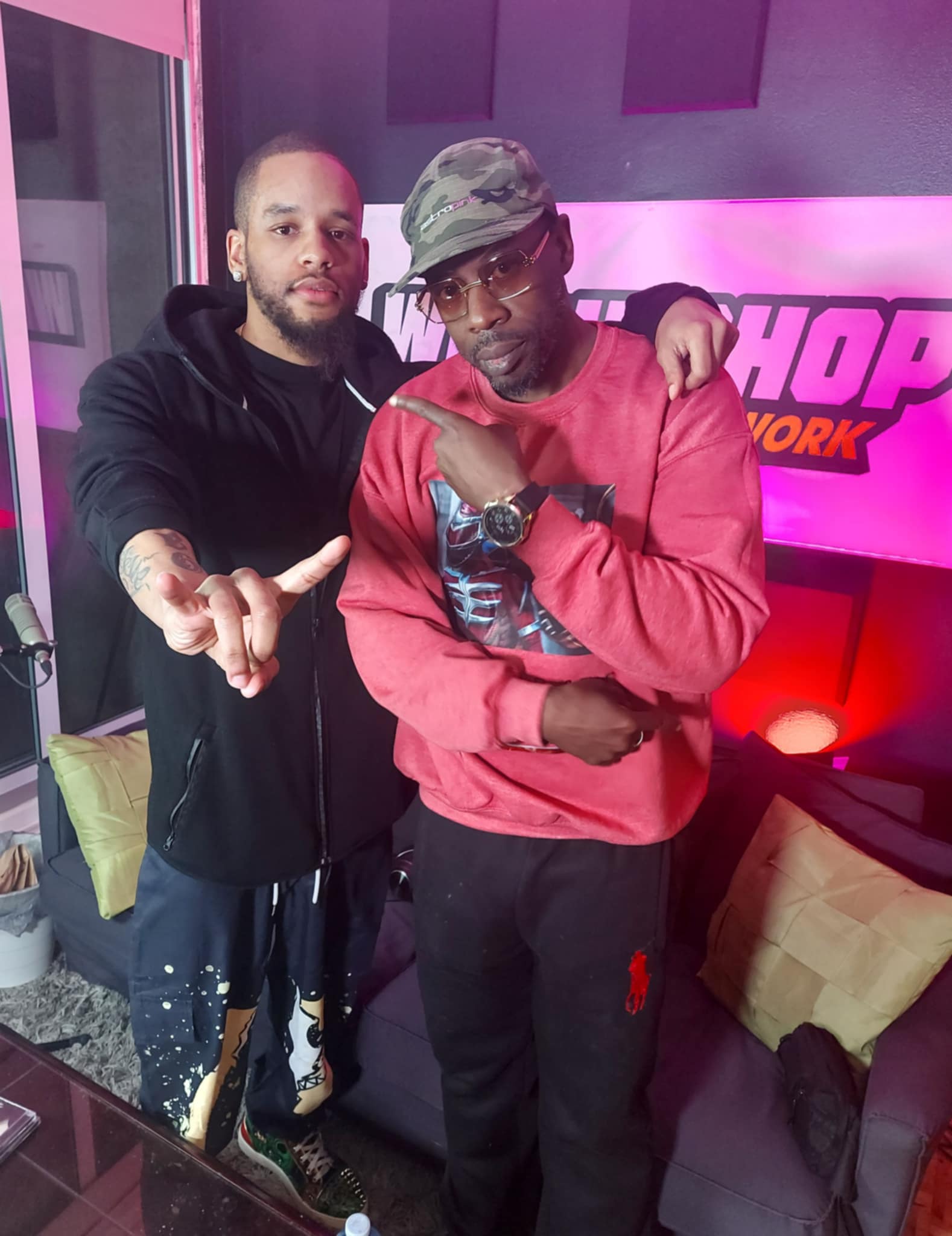 Two men pose together in a room with purple and pink lighting, one wearing a black hoodie and the other a red sweatshirt and camouflage cap.