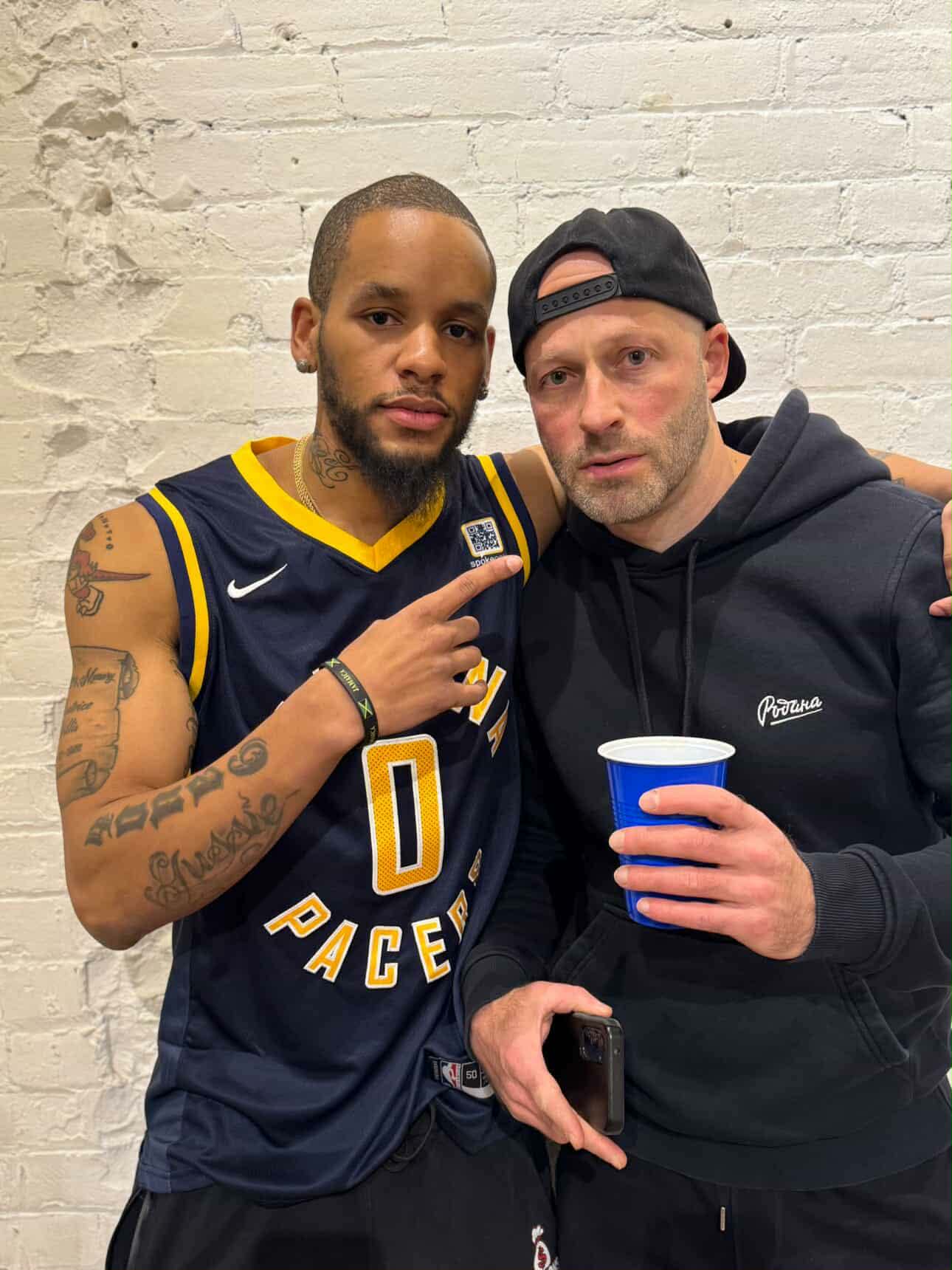 Two men pose together against a white brick wall, one wearing a basketball jersey and the other in a hoodie, holding a blue cup.