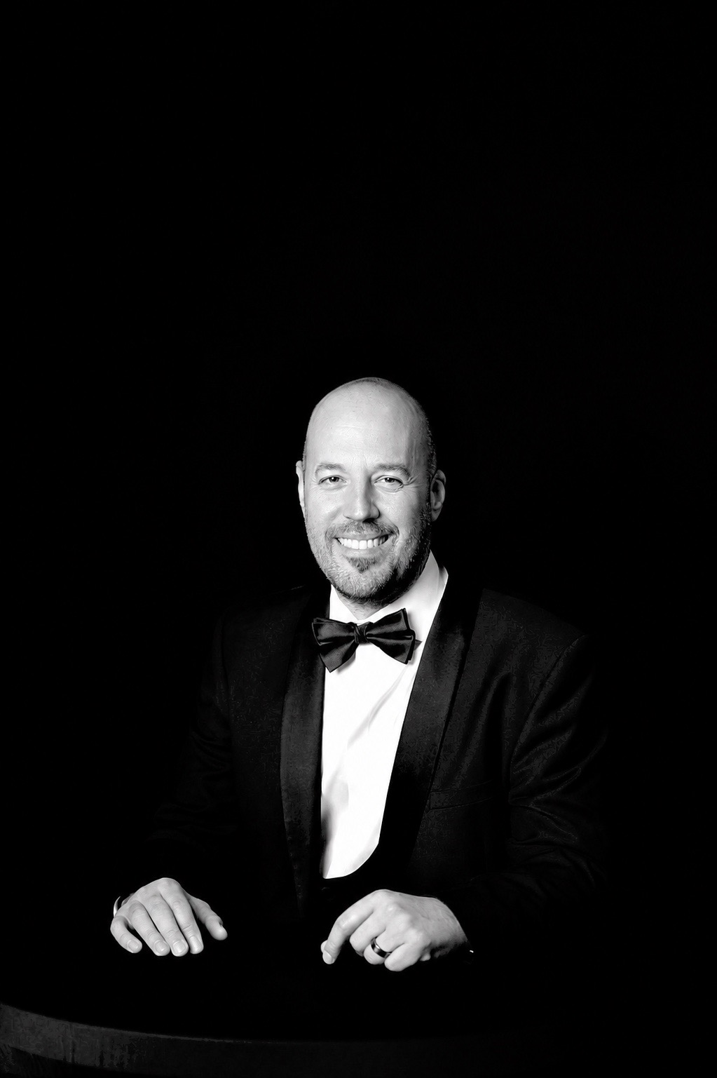 Man in tuxedo with bow tie smiling, seated against a black background.